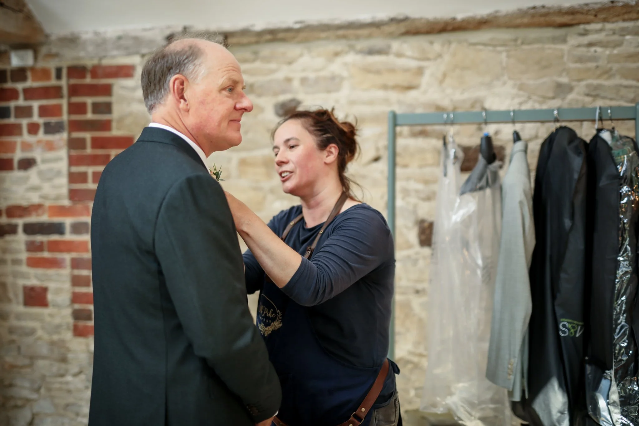 A man in a tuxedo and a woman in a dark dress are in a rustic room with exposed brick and stone walls. The woman is pinning a boutonniere on the man's lapel, and they are smiling at each other.