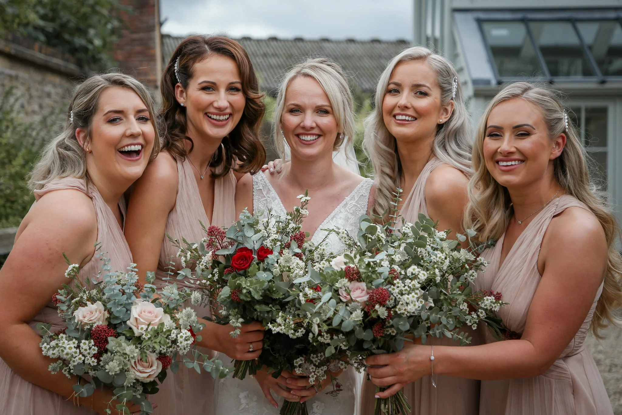 A bride and five bridesmaids smiling and holding bouquets of flowers outdoors during a wedding celebration.