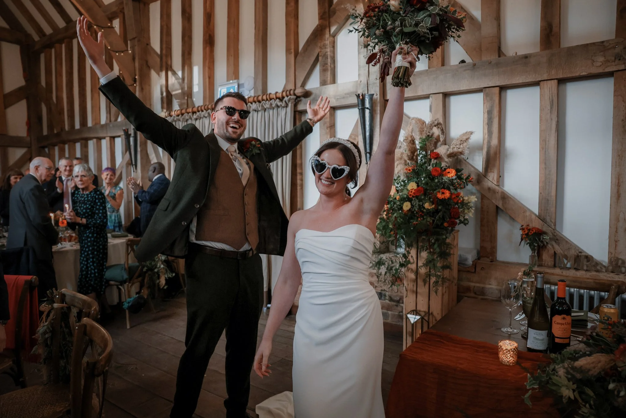 Man and woman celebrating at a wedding reception, wearing sunglasses; the woman is holding a bouquet and the man has his arms raised in celebration, with guests clapping in the background.