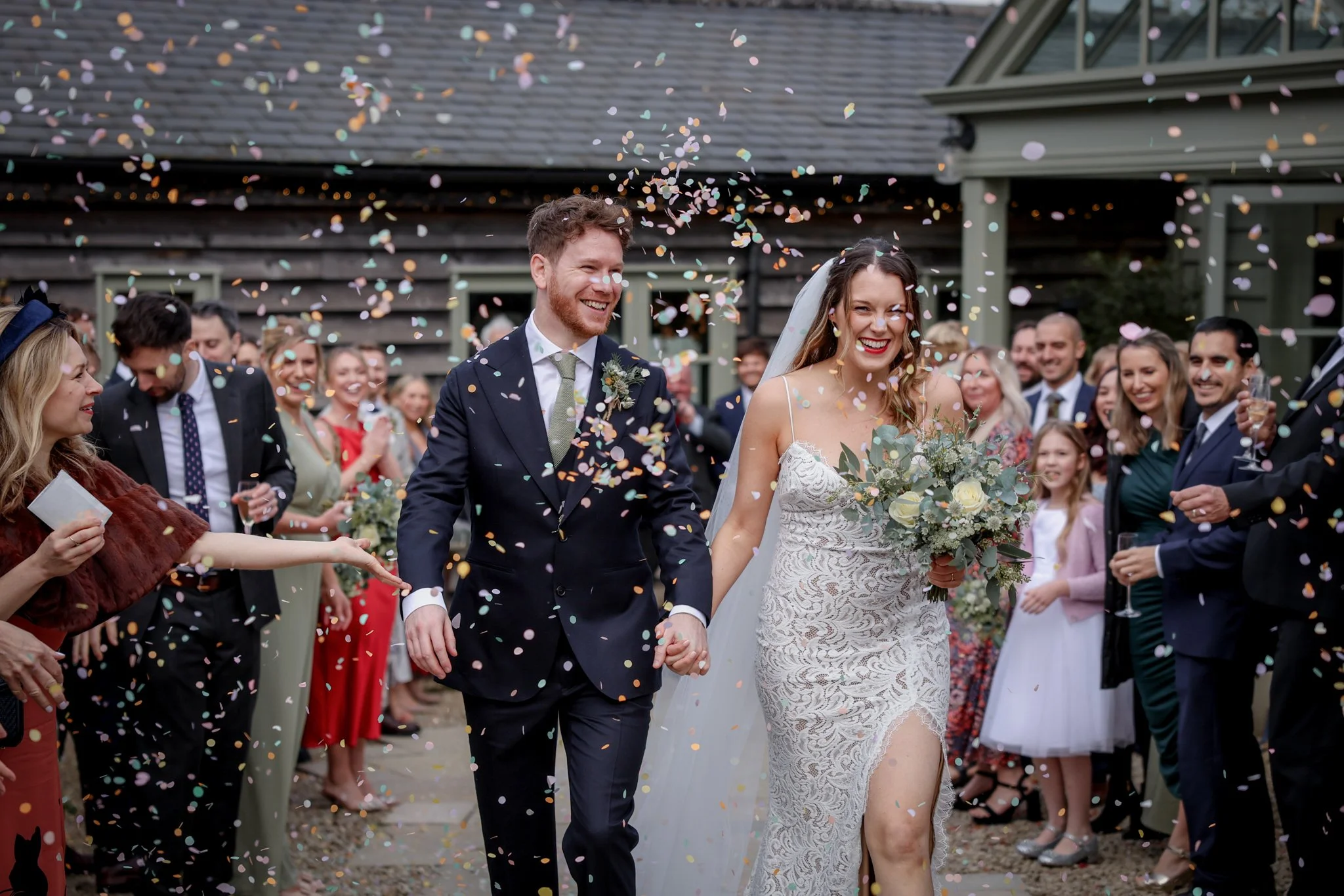 A bride and groom smiling and holding hands, walking through a crowd of celebrating guests, while colorful confetti falls around them during a wedding celebration outside.