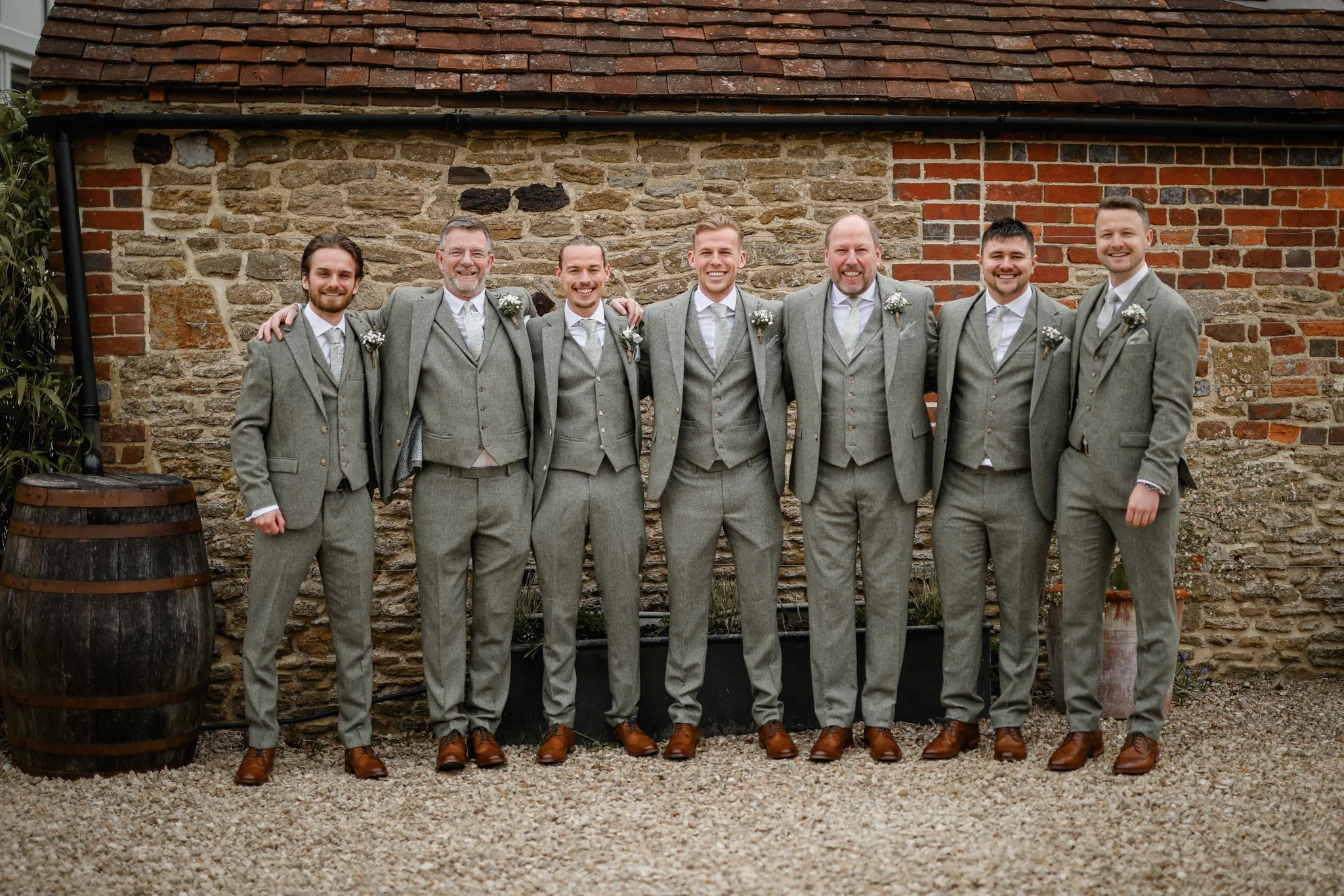 Seven men dressed in matching gray suits and brown shoes standing together outdoors in front of a brick and stone wall, smiling with arms around each other.