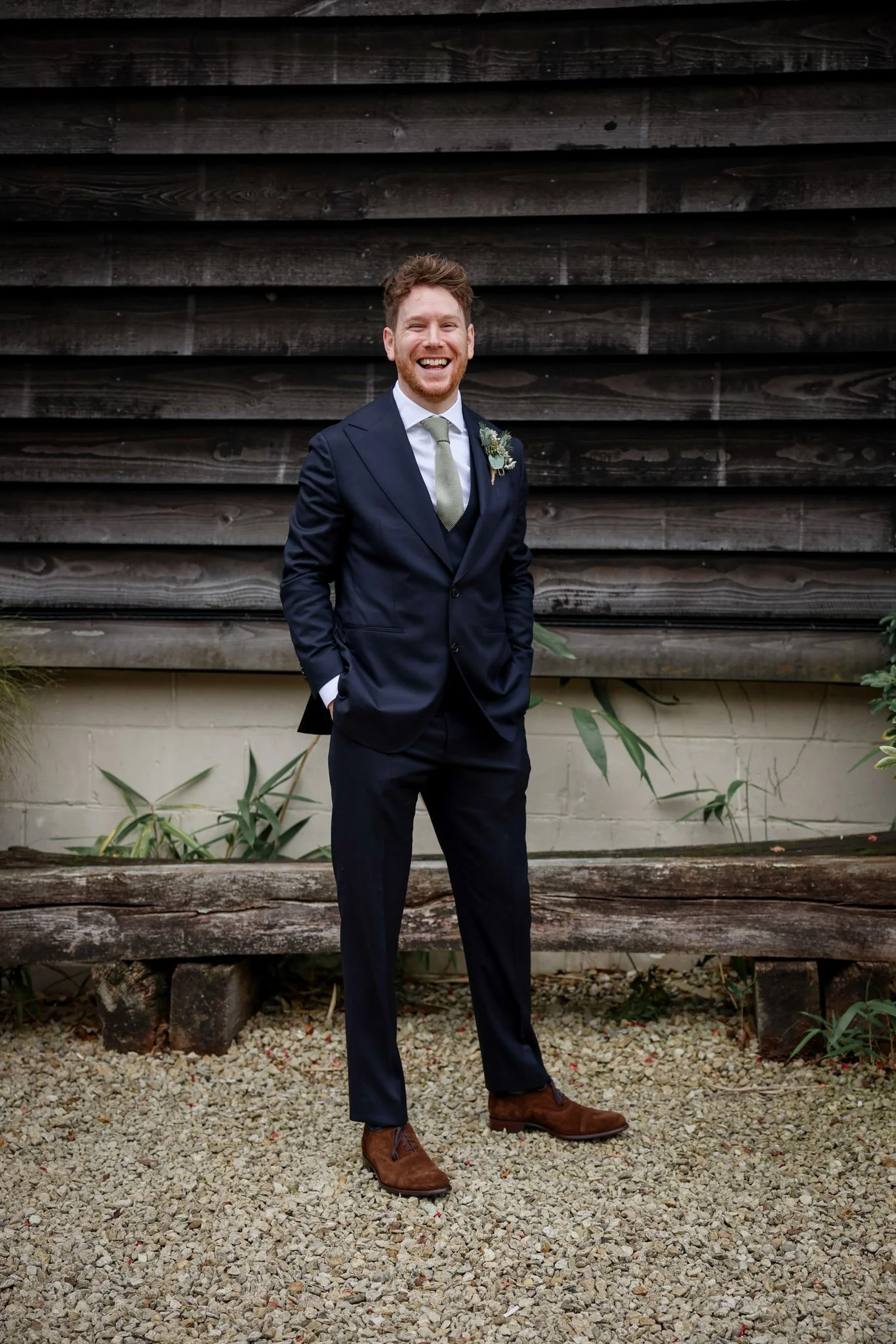 A man in a navy suit and green tie standing outdoors on a gravel surface, smiling broadly, with a wooden fence behind him.