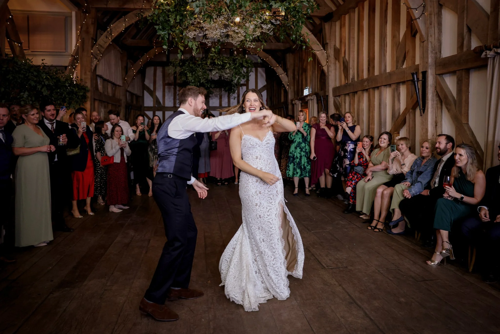 A wedding reception with a bride and groom dancing while friends and family watch and take photos in a rustic barn with string lights and floral decorations.