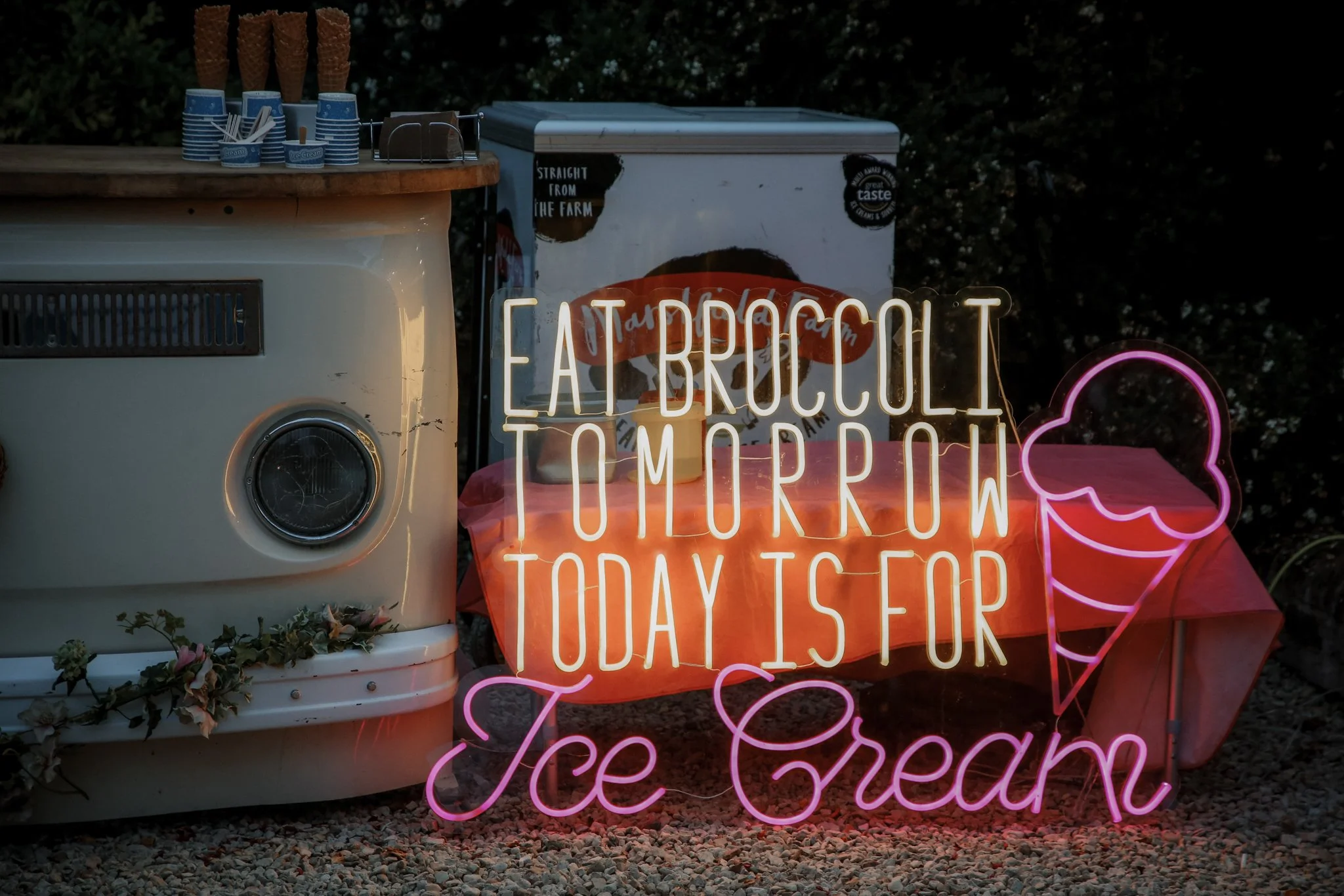 Ice cream stand with a neon sign that reads 'Farmhouse tomorrow today stop for ice cream,' positioned next to a vintage ice cream freezer and a table with ice cream cones and cups. The scene is set outdoors in the evening.