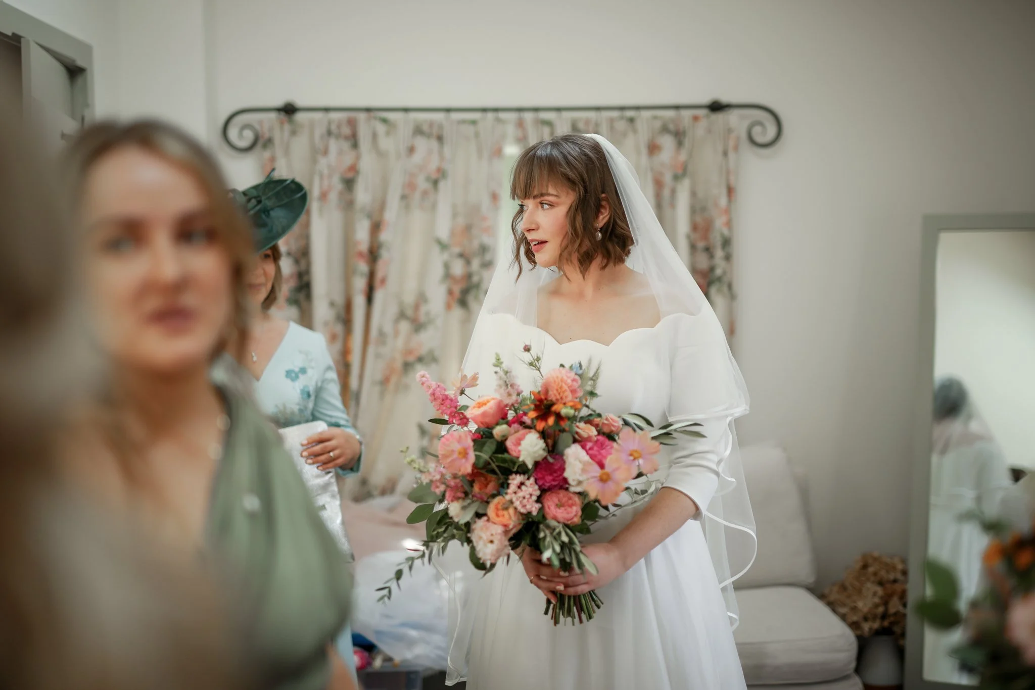 A bride in a white dress and veil holding a large bouquet of pink, peach, and white flowers, preparing for her wedding surrounded by women, with a floral curtain in the background.