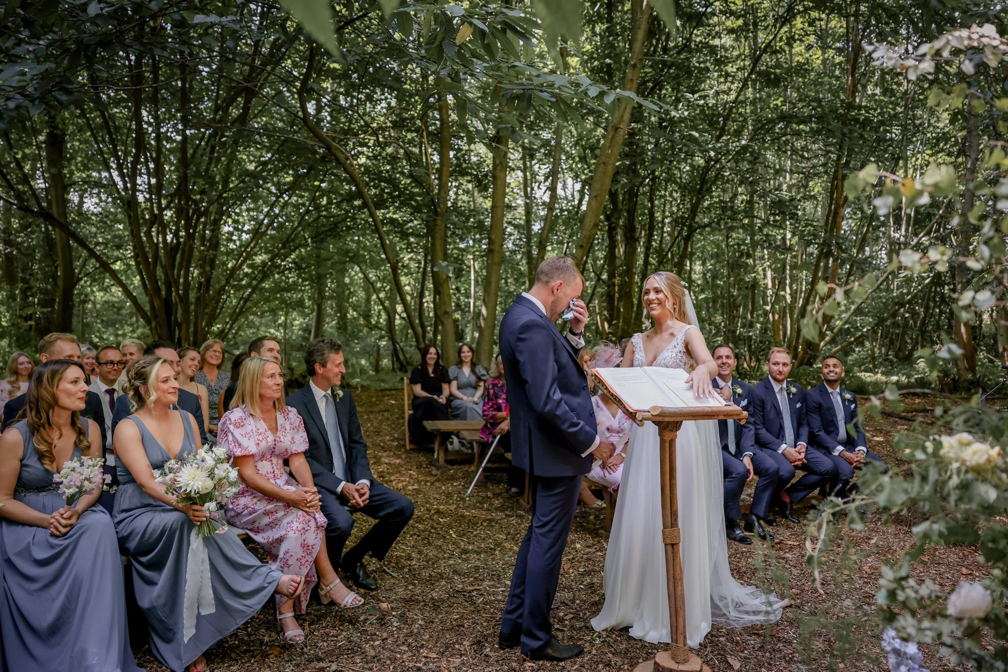 A couple gets married in an outdoor woodland setting surrounded by friends and family, with the groom wiping away tears and the bride smiling during the ceremony.