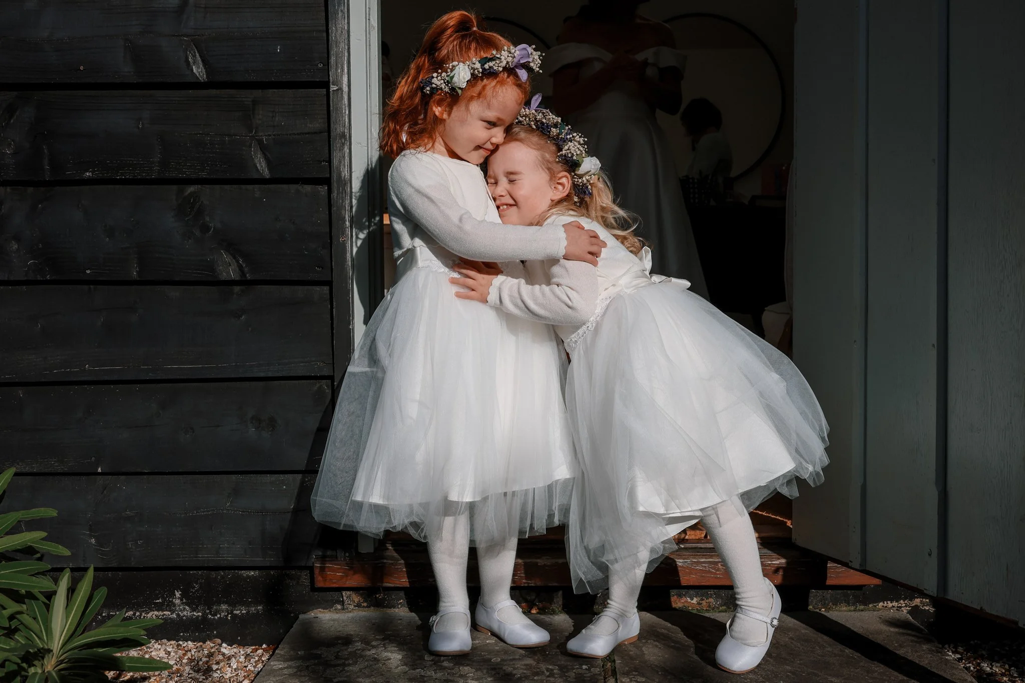 Two young girls wearing white dresses and flower crowns hugging each other outside near a dark wooden wall and a light-colored building.