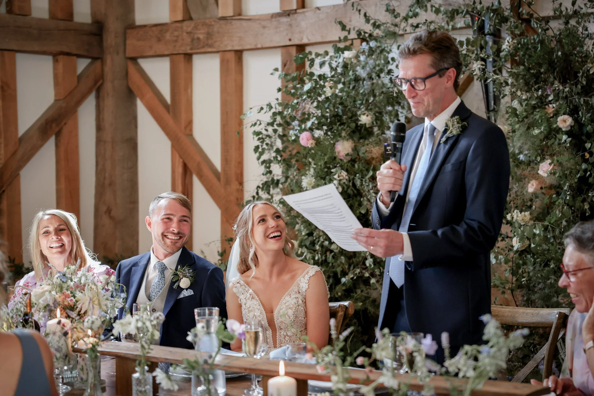 A man in a suit and glasses is giving a speech at a wedding reception, standing in front of a floral backdrop, with the bride and groom and guests sitting at a decorated table in a rustic barn setting.