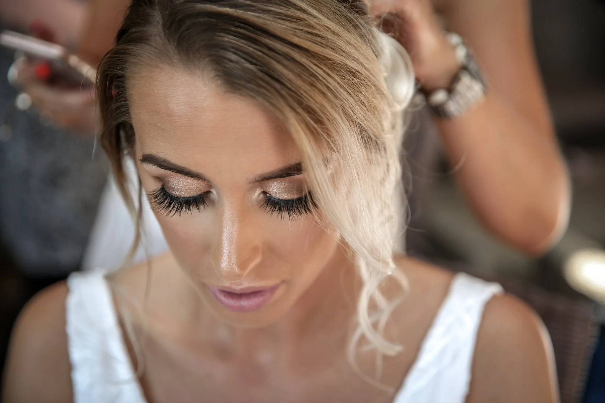 Close-up of a woman with styled blonde hair and long eyelashes, looking downward, wearing a white top.