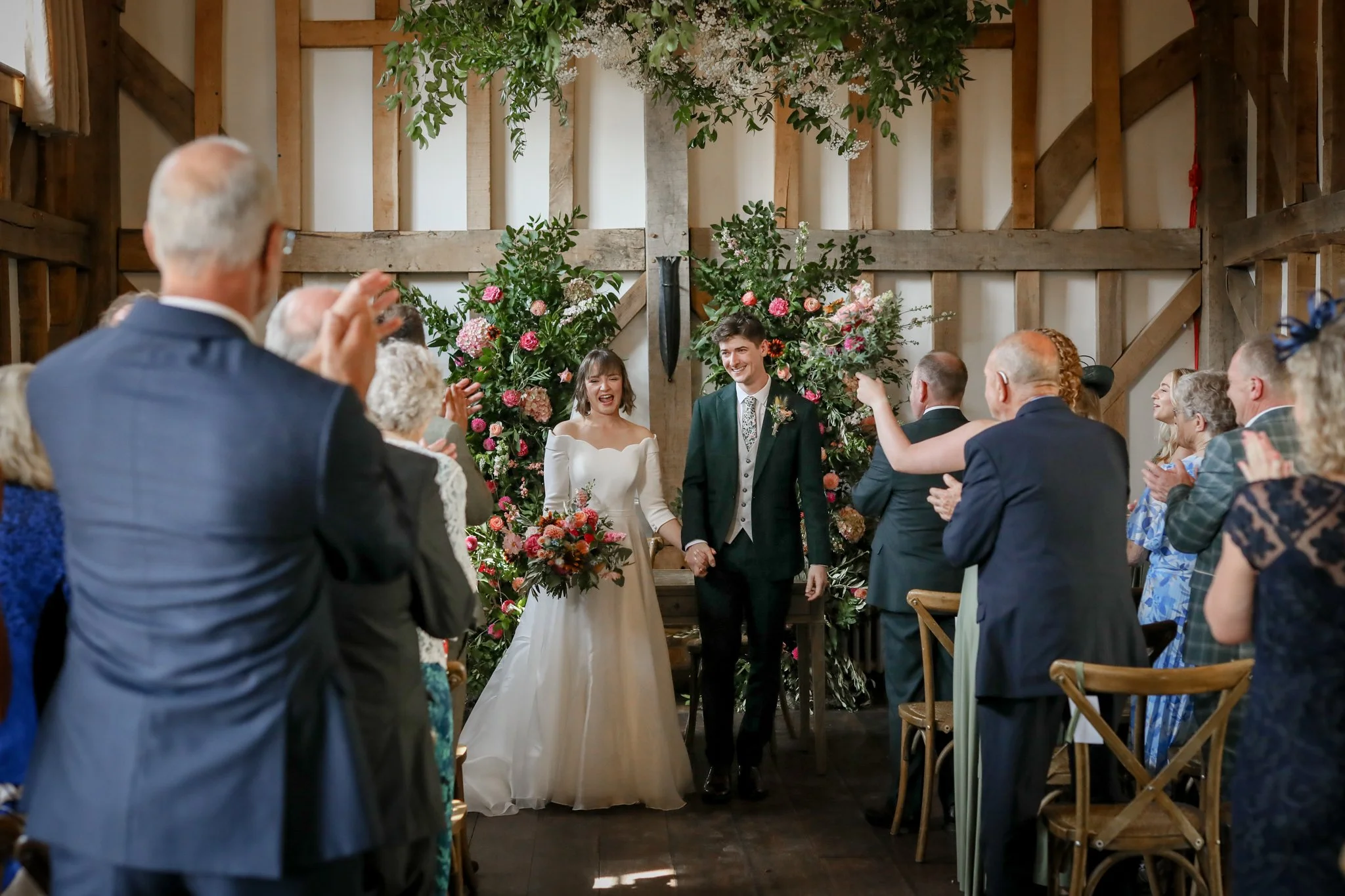 A wedding ceremony with a bride and groom standing in front of an altar adorned with colorful flowers. Guests are seated on wooden chairs, some clapping and smiling, inside a rustic venue with wooden beams and floral decorations hanging from the ceil
