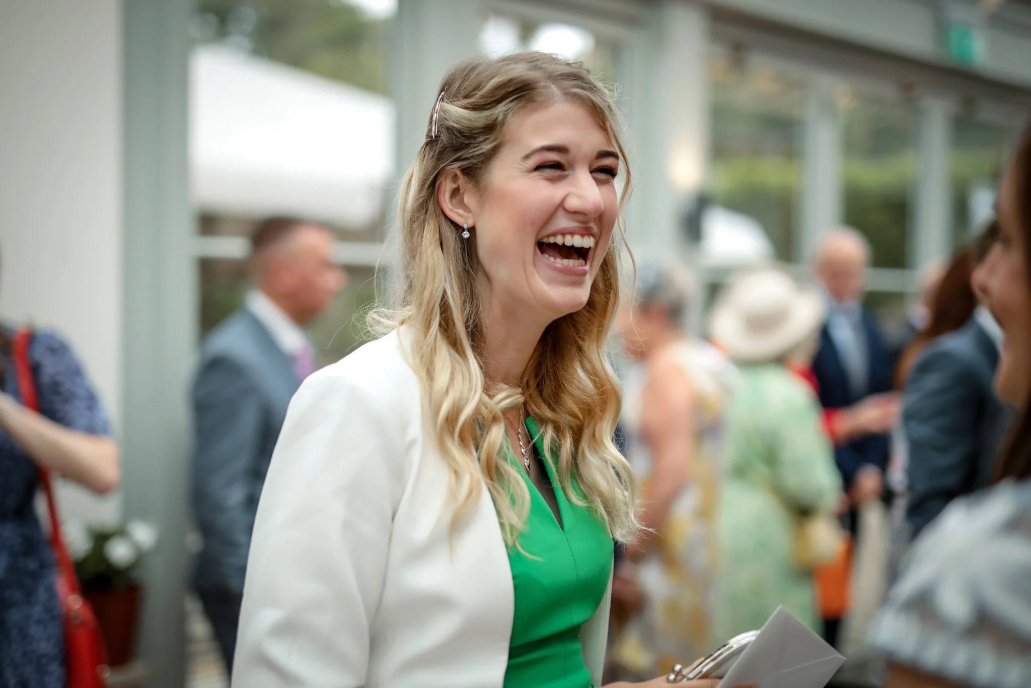 A woman with long blonde hair smiling while talking to someone at an indoor event, with others blurred in the background.