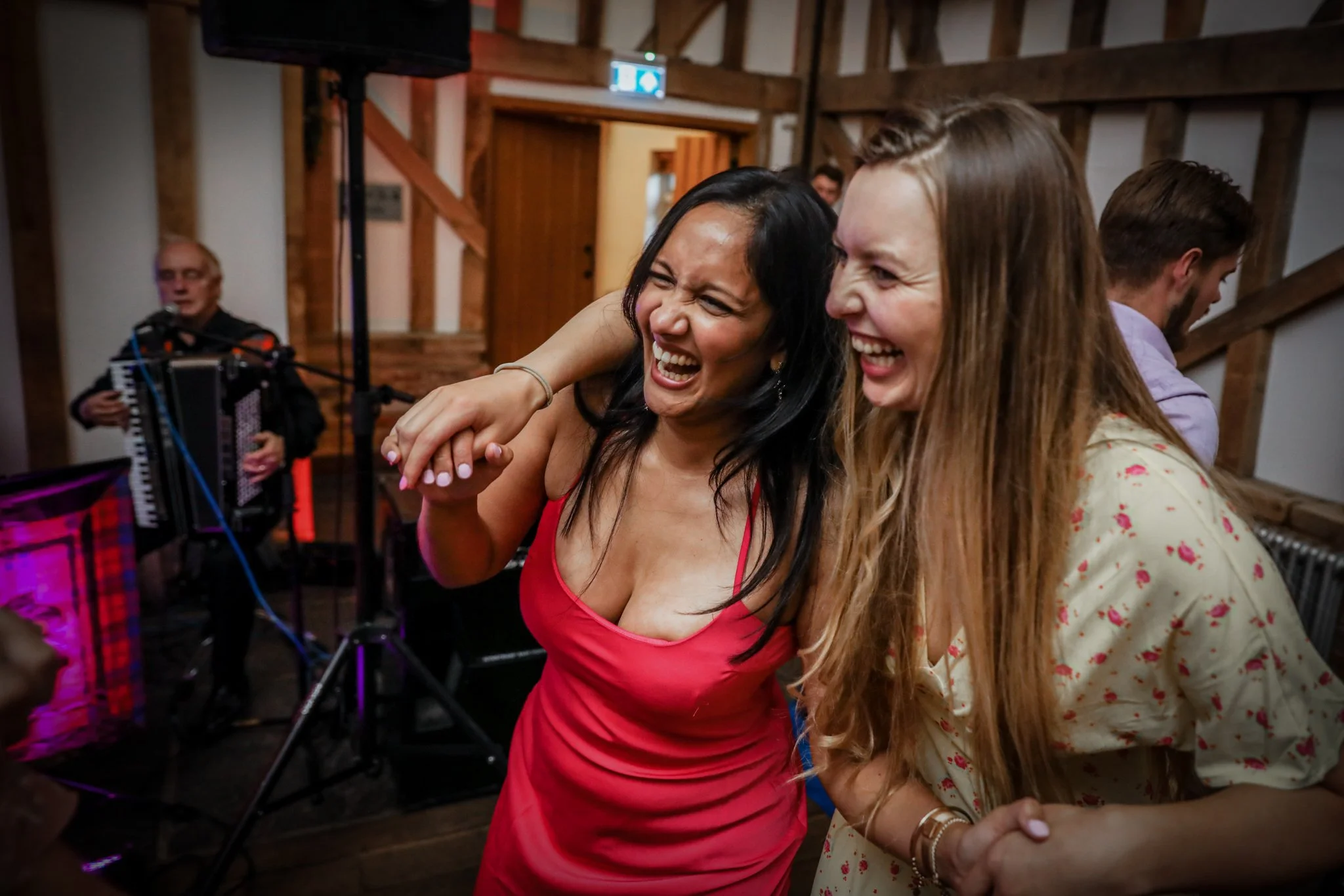 Two women dancing closely together, smiling and laughing, at a social event with a musician playing an accordion in the background.
