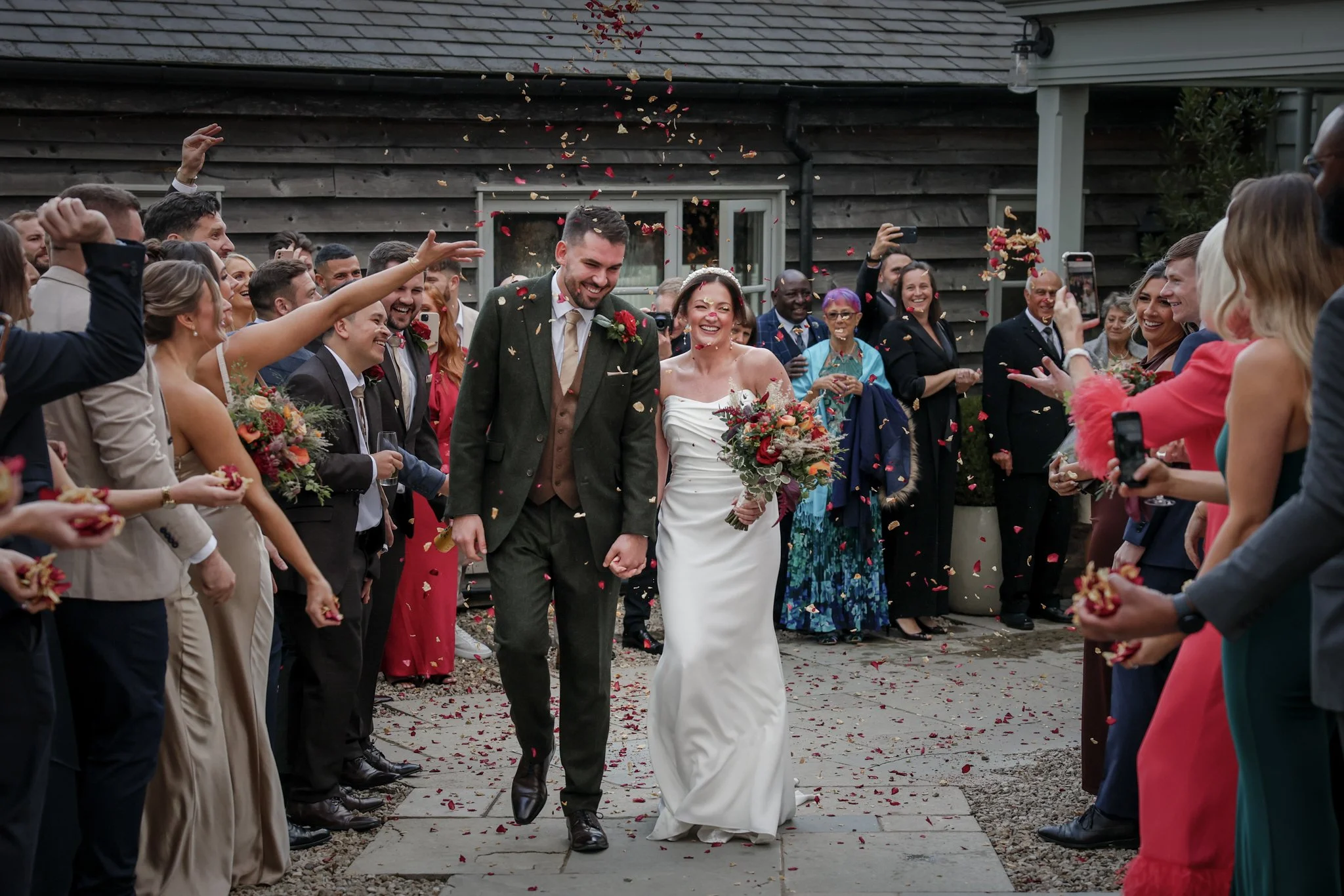A newly married couple walking down a path, smiling, while guests throw flower petals in celebration at an outdoor wedding.