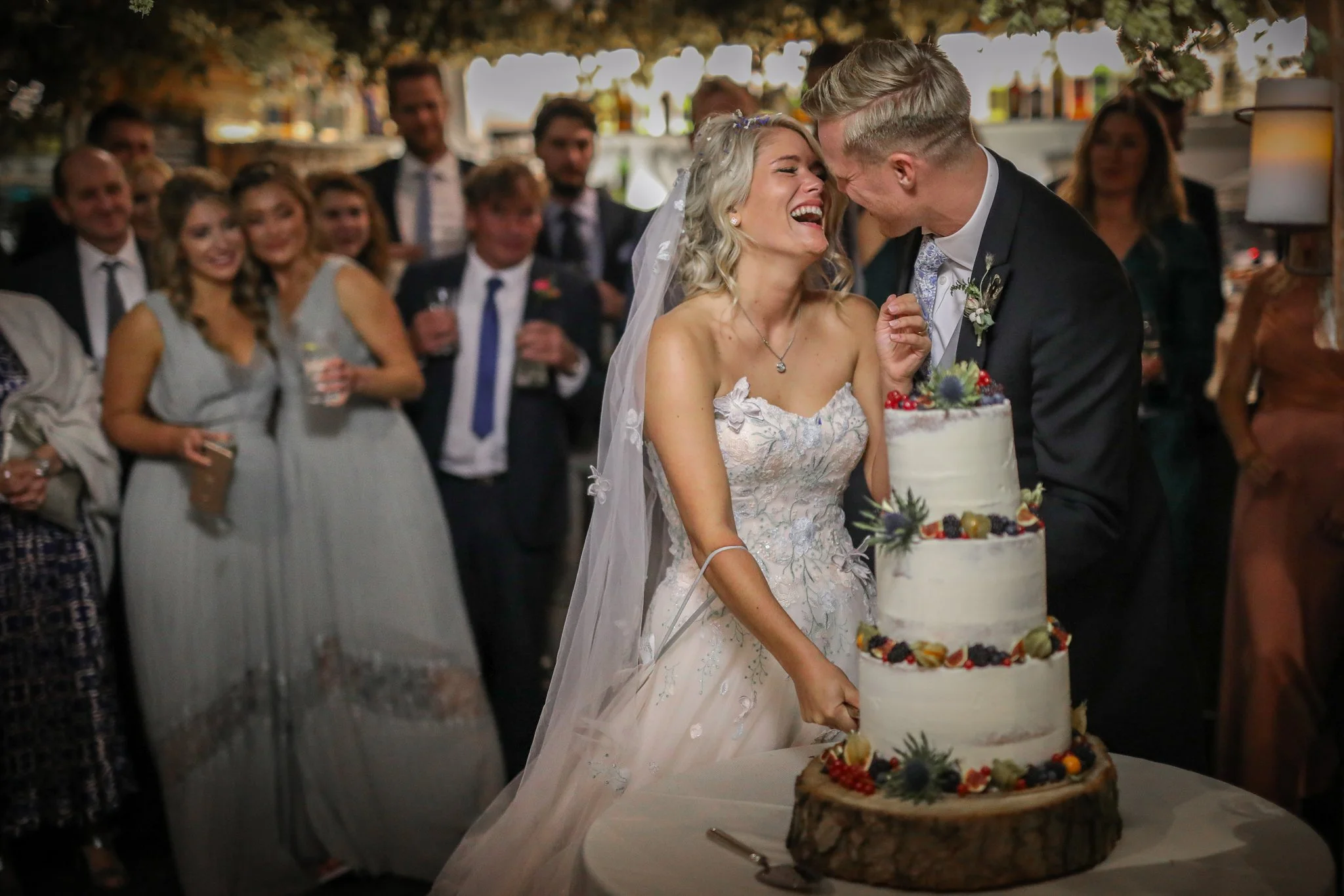 A bride and groom smile and laugh as they prepare to cut their wedding cake at a reception, surrounded by wedding guests in an outdoor setting.