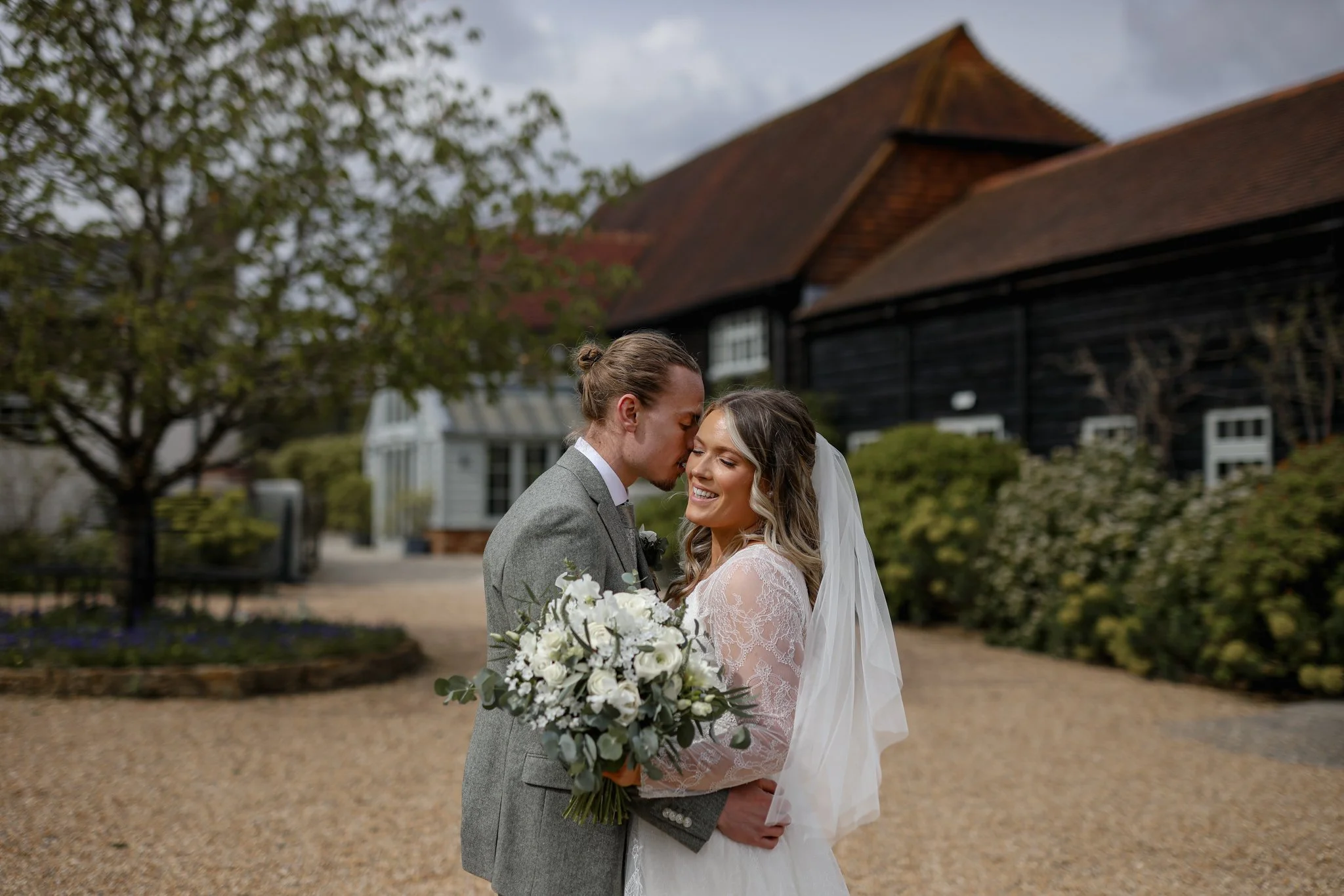 An interracial bride and groom kissing outdoors during their wedding, with the bride holding a bouquet of white flowers, and buildings with pagoda-style roofs in the background.