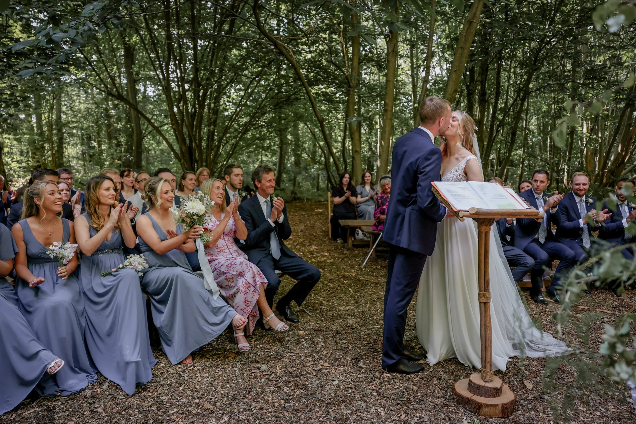 A couple shares a kiss during their outdoor wedding ceremony in a forest, surrounded by seated guests in formal attire, some holding flowers, with greenery and trees in the background.