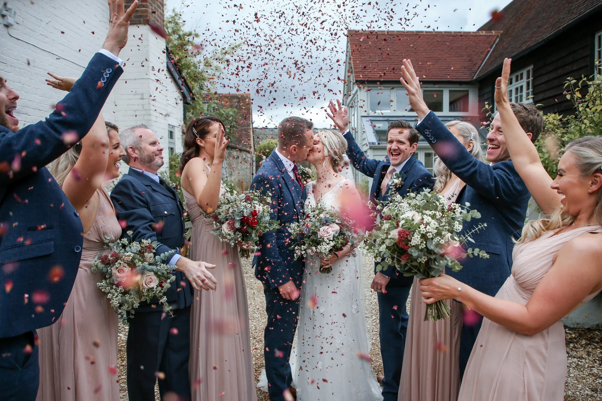 A wedding celebration outdoors with a group of people throwing flower petals and confetti in the air as the bride and groom kiss during their ceremony, surrounded by friends and family.