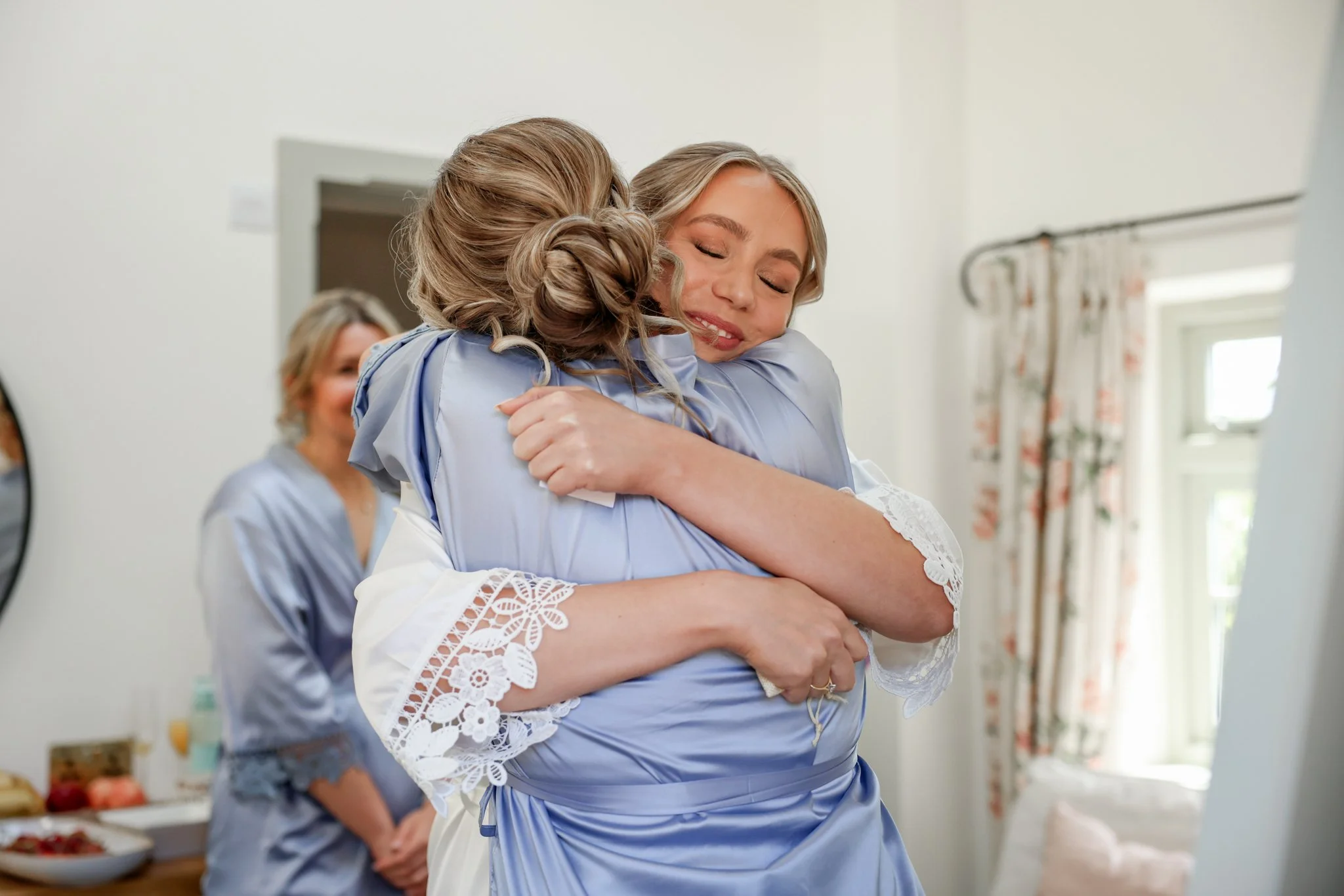 Two women sharing a hug indoors, with a woman in the background near a kitchen counter with fruit and bowls.