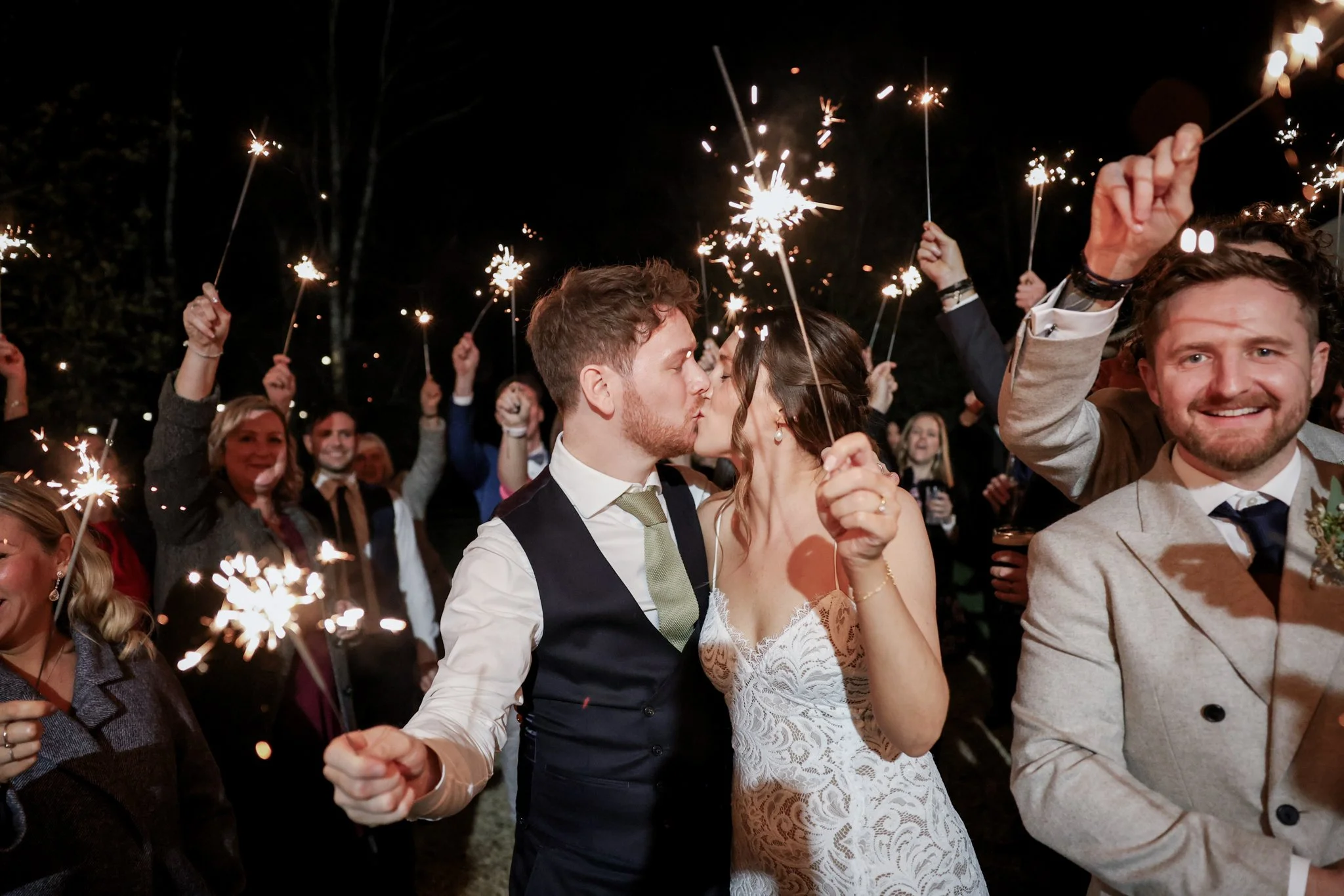 A newlywed couple kisses at night during a celebration with sparklers, surrounded by friends and family.