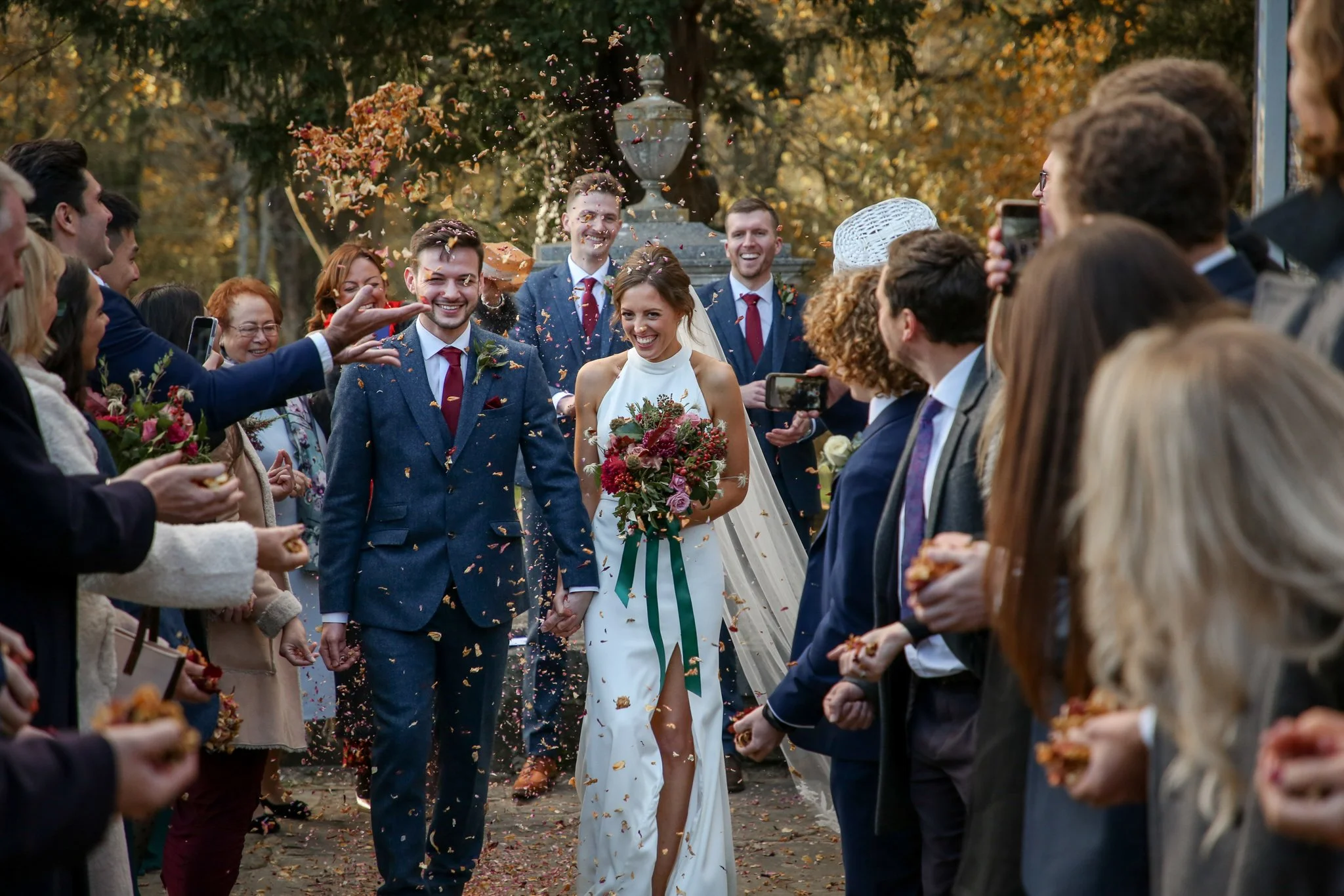 A bride and groom holding hands while walking through an outdoor wedding celebration, surrounded by family and friends, with confetti falling around them.