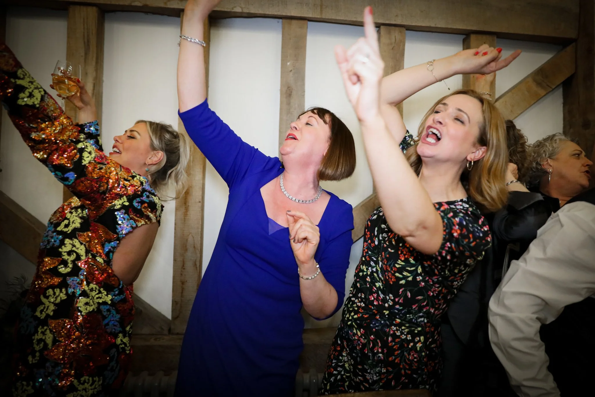 Group of women dancing and celebrating at an indoor gathering, smiling, with one holding a glass of wine, under wooden beams.