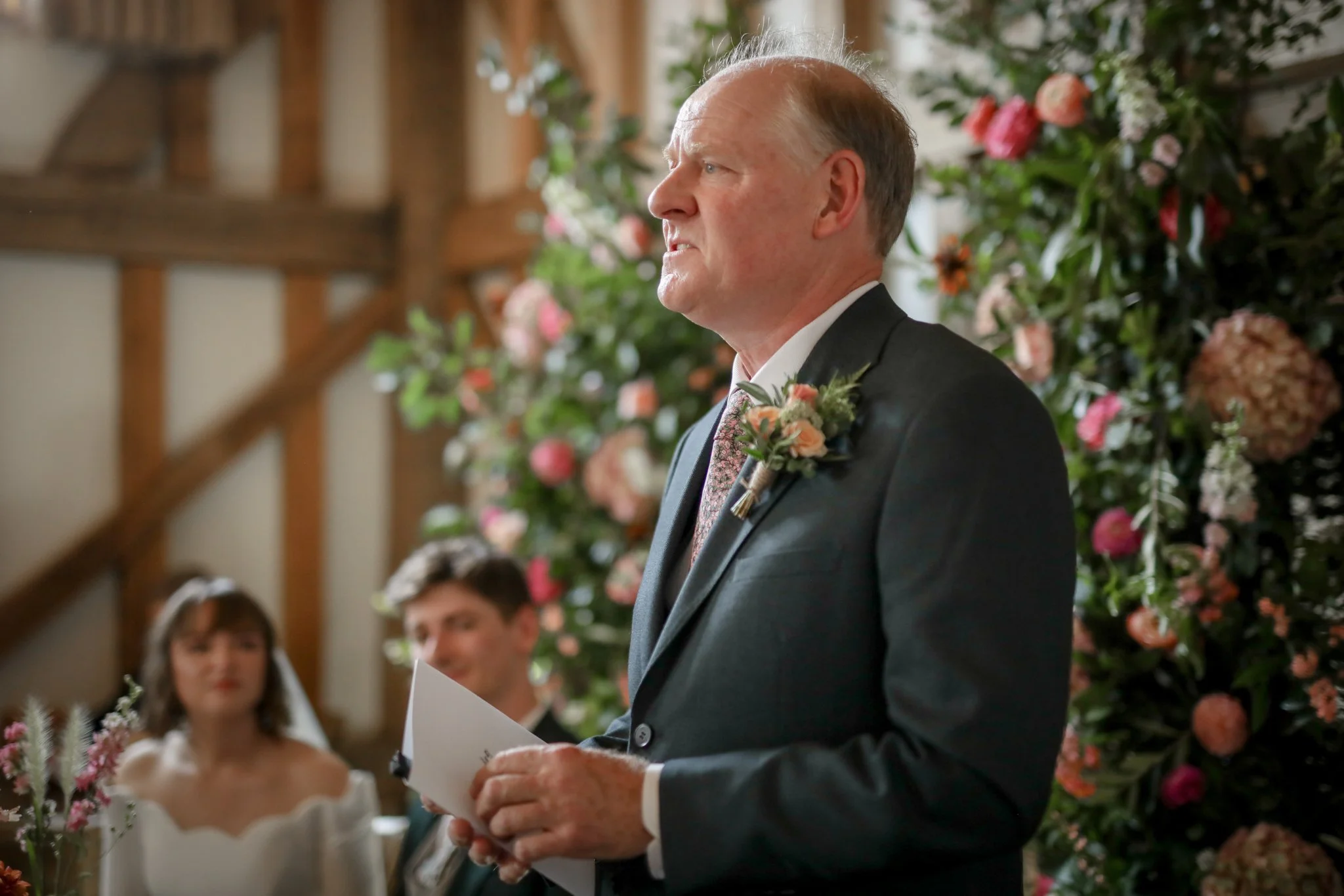 An elderly man in a black suit and floral tie, with a boutonniere, is standing in front of a floral backdrop, holding a piece of paper, during a wedding ceremony.