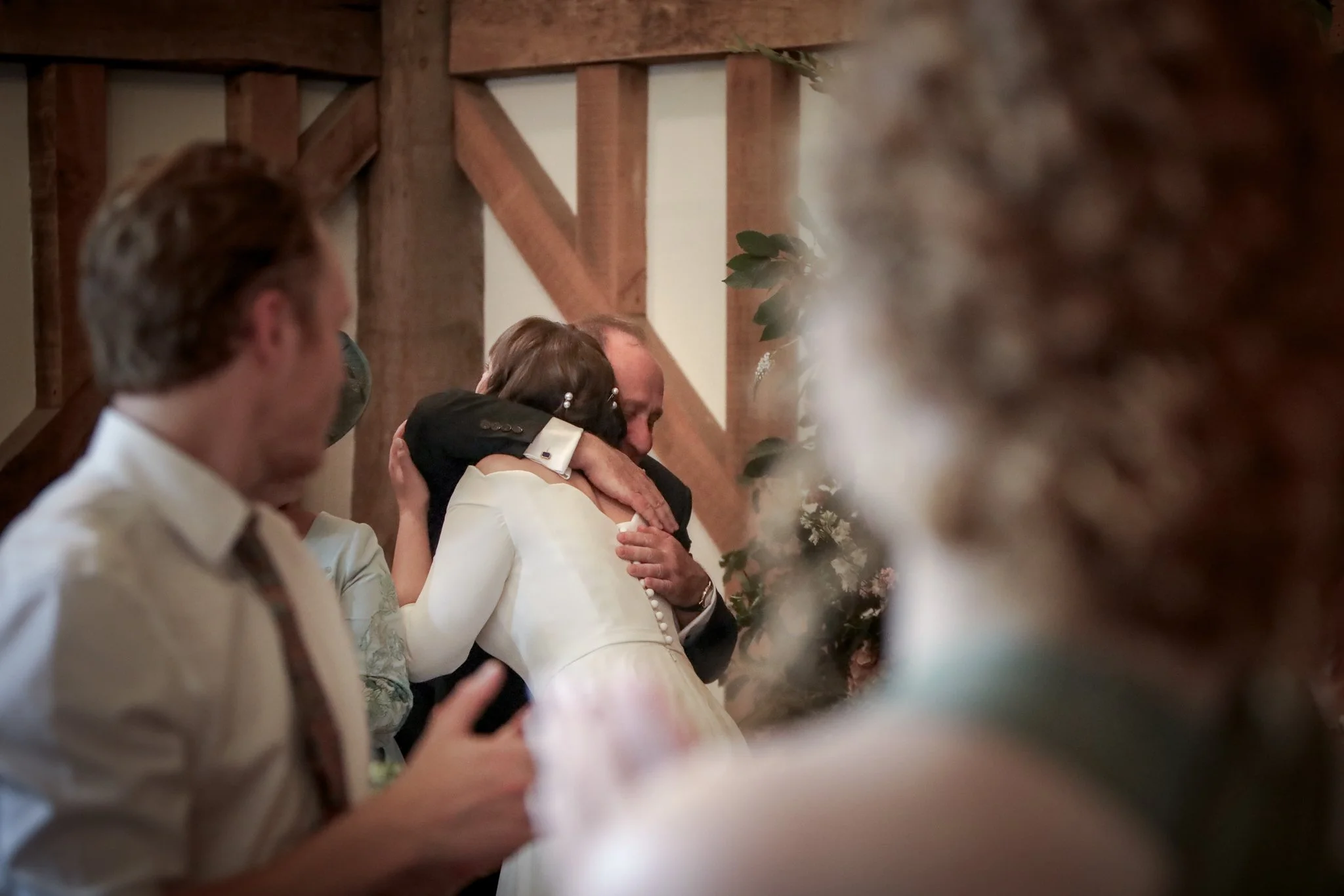 A bride and groom sharing a hug during a wedding ceremony, with wedding guests in the foreground and a wooden wall with greenery in the background.