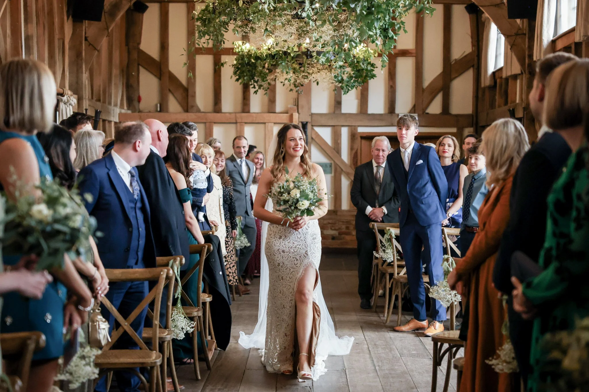 A bride in a lace wedding dress walking down the aisle in a rustic wooden barn decorated with hanging greenery and flowers, surrounded by seated guests.