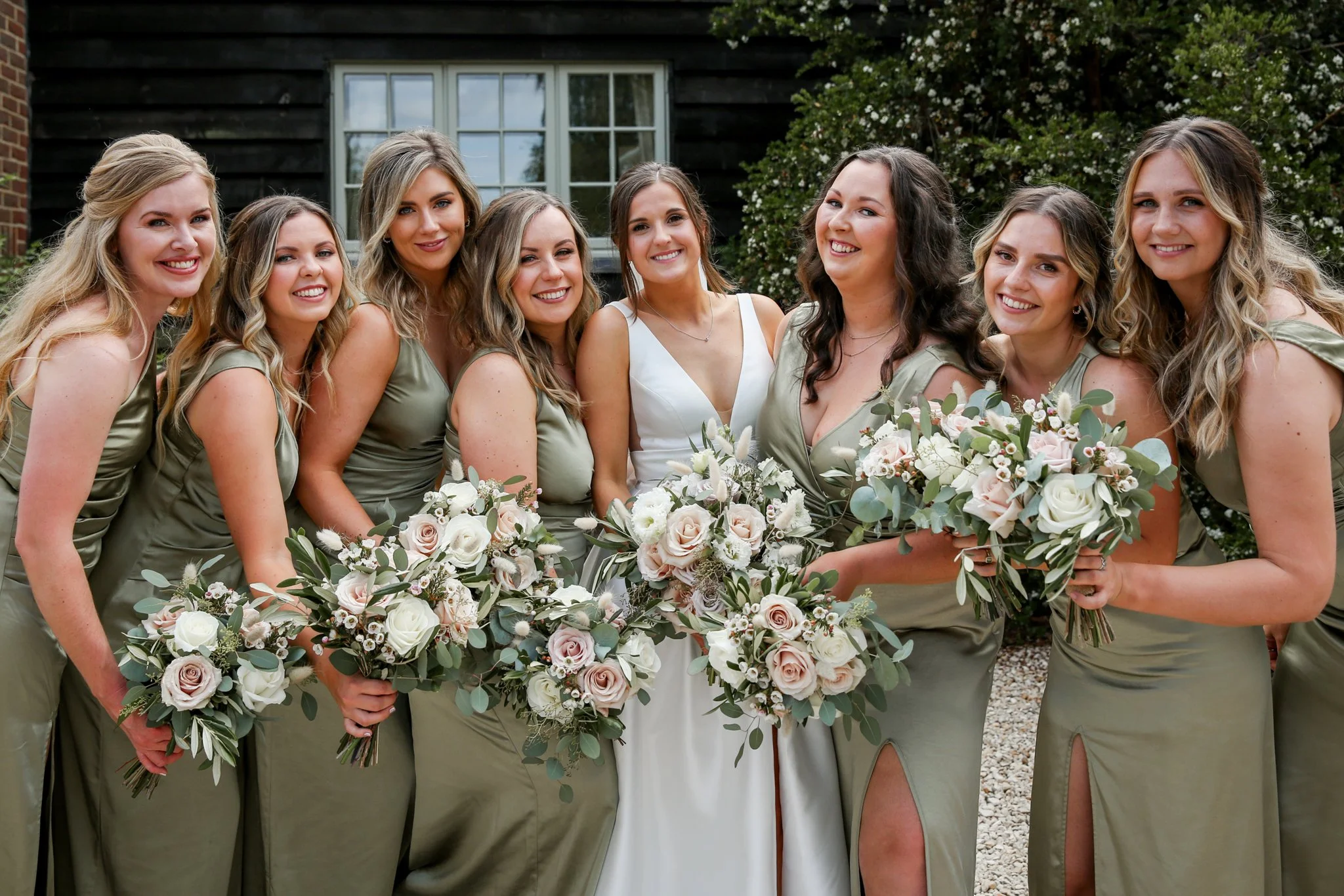 Group of women dressed in matching green dresses, holding bouquets, standing outdoors near a black building with white windows and greenery.