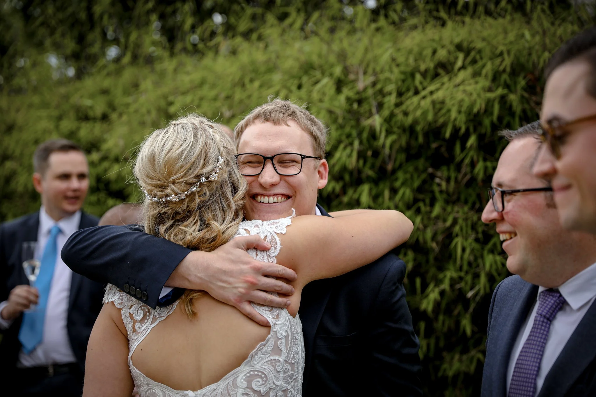 Bride and groom hugging during wedding celebration outdoors, with other guests in background.
