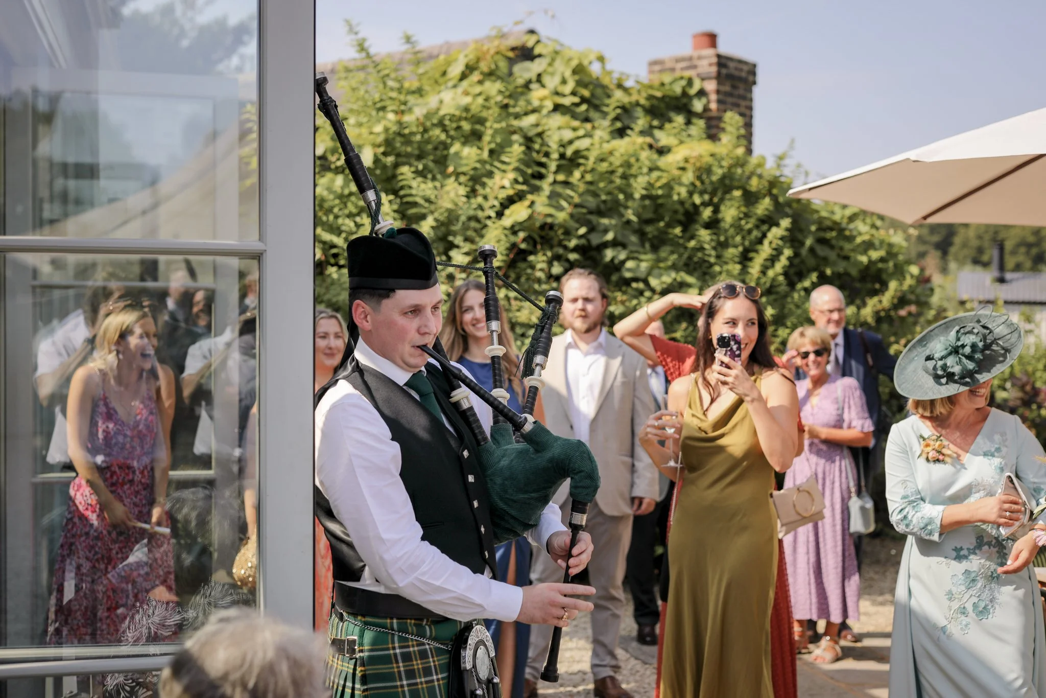 A man playing bagpipes at an outdoor gathering, with a group of people watching and smiling behind him, during a sunny day with trees and a building in the background.