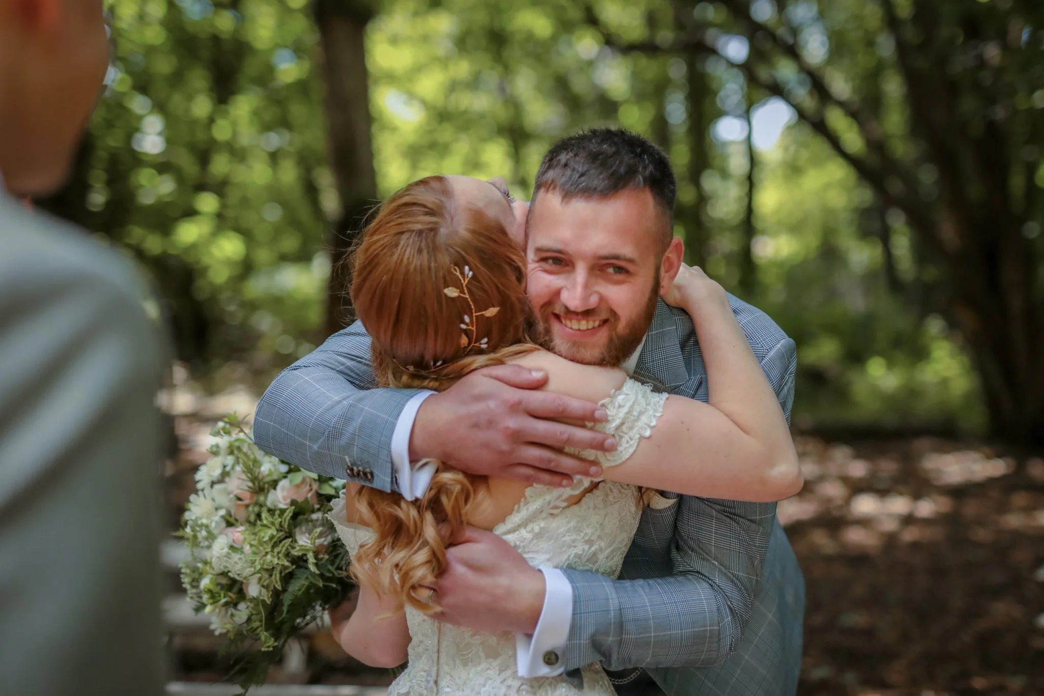 A man in a gray suit embraces a woman in a white dress with lace details, likely at a wedding or romantic event, in an outdoor setting with trees and sunlight in the background.