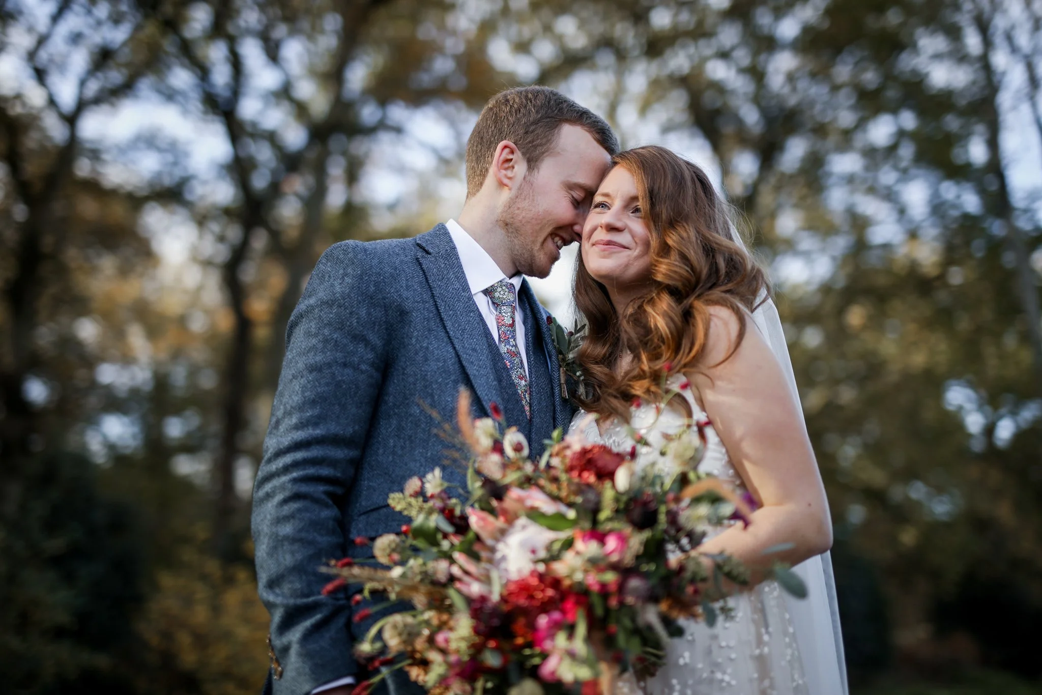 A happy couple, a man and a woman, close together with their foreheads touching, smiling outdoors during daytime. The man is wearing a gray suit jacket, and the woman has long, wavy brown hair and is holding a colorful bouquet of flowers.
