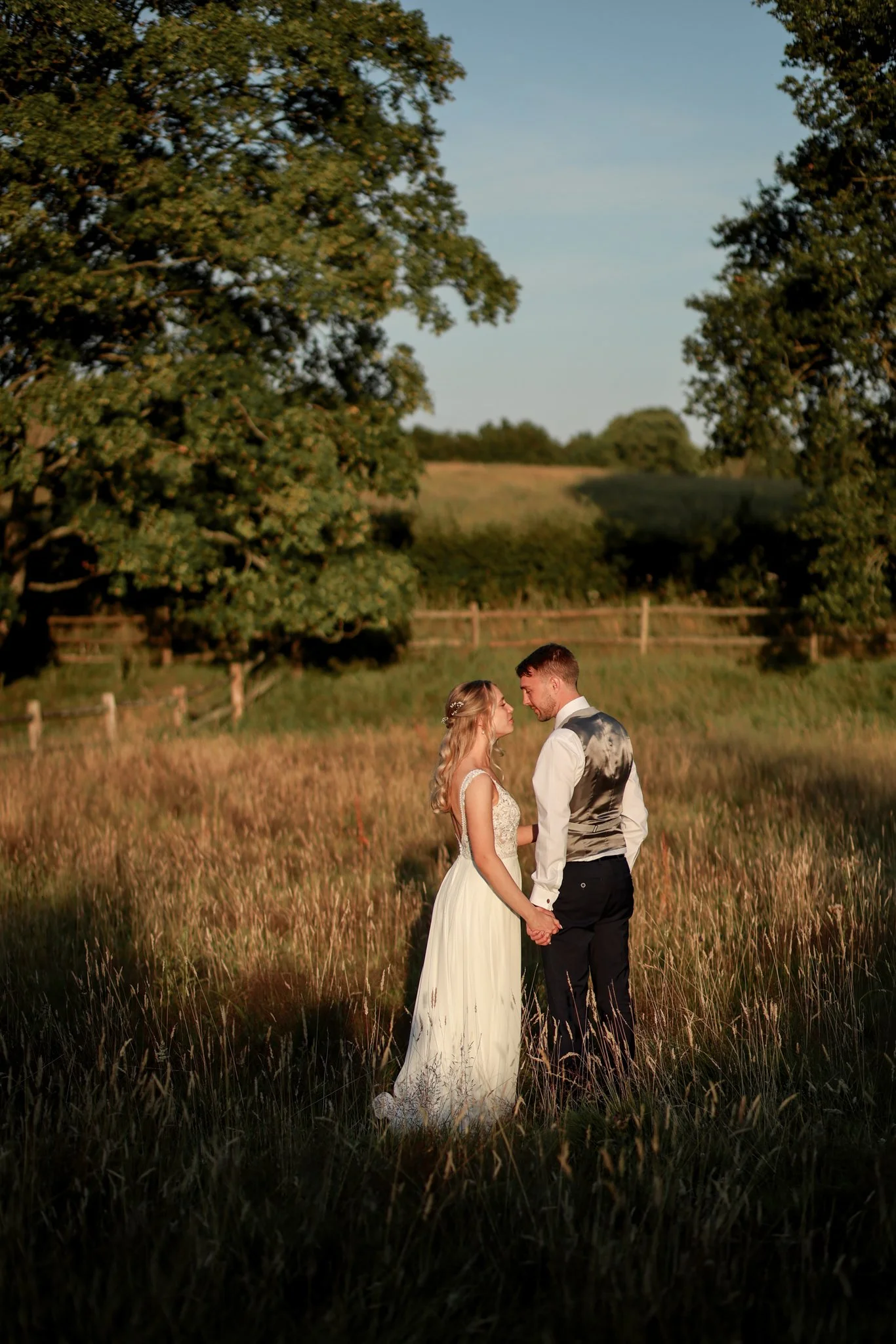 A bride and groom holding hands and looking into each other's eyes in a field with tall grass, trees, and a fence in the background, during sunset.