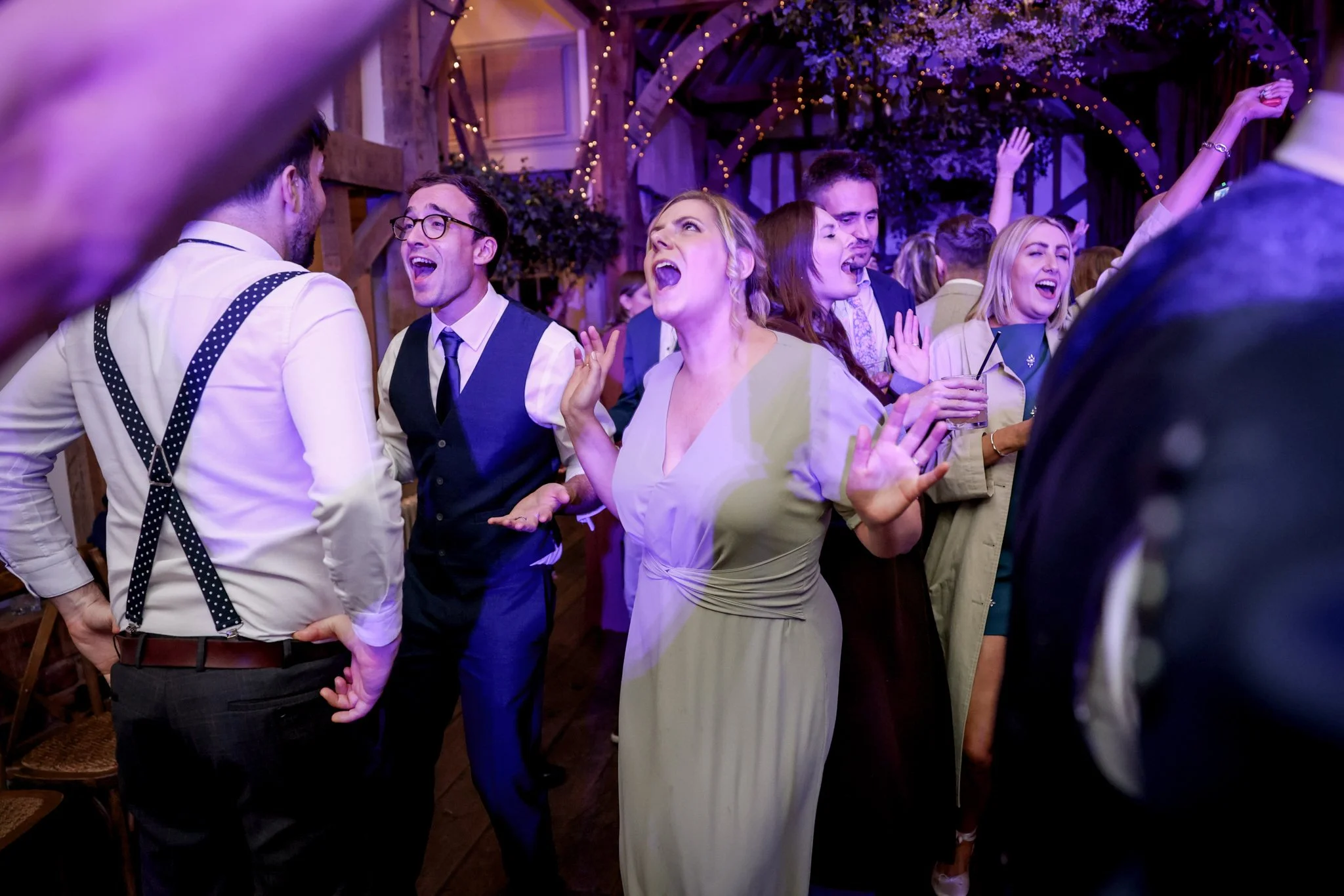 People dancing and singing at a lively indoor celebration with string lights and purple lighting.