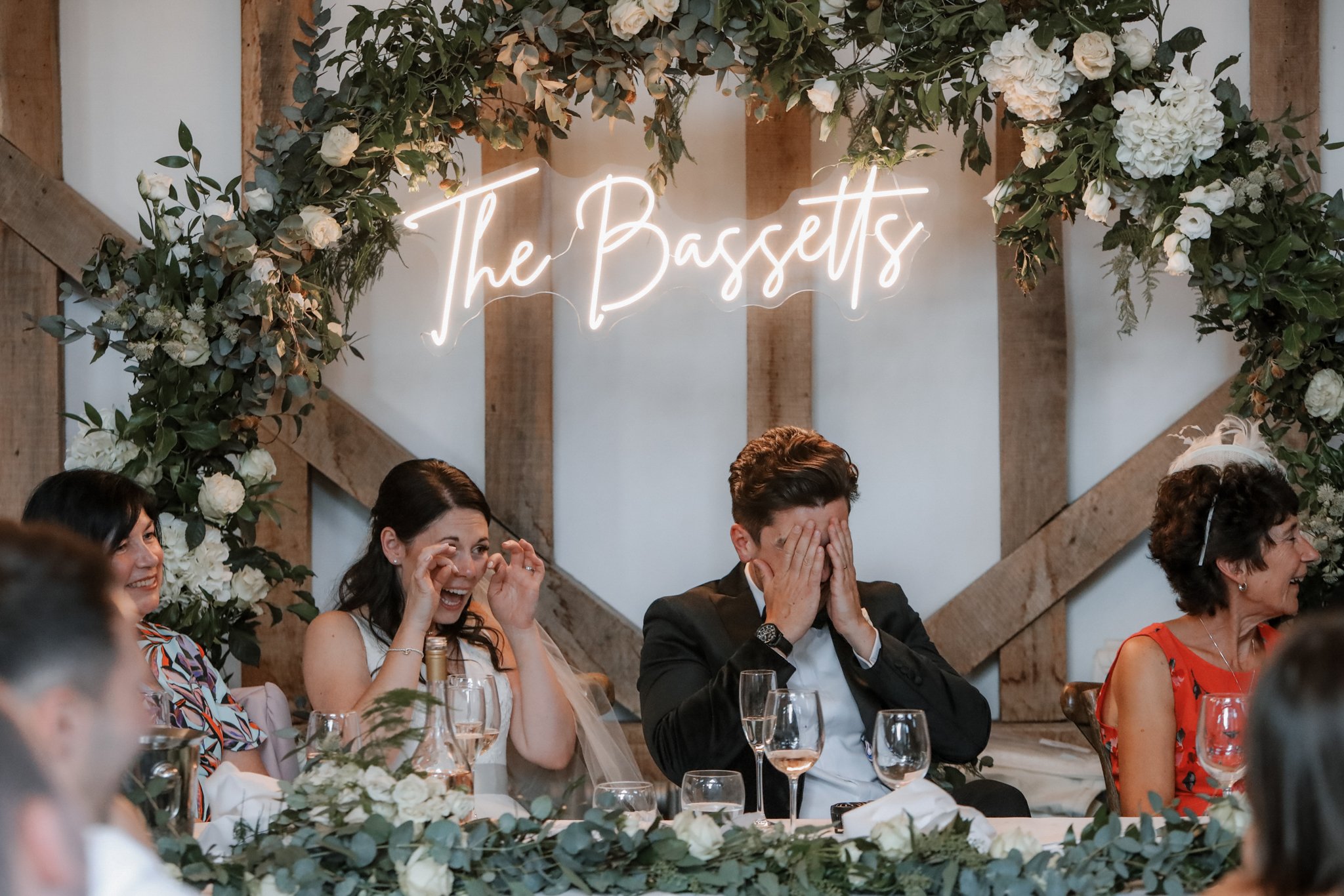 People sitting at a decorated wedding reception table, smiling and reacting emotionally, with a floral arch and neon sign reading 'The Bassets' in the background.