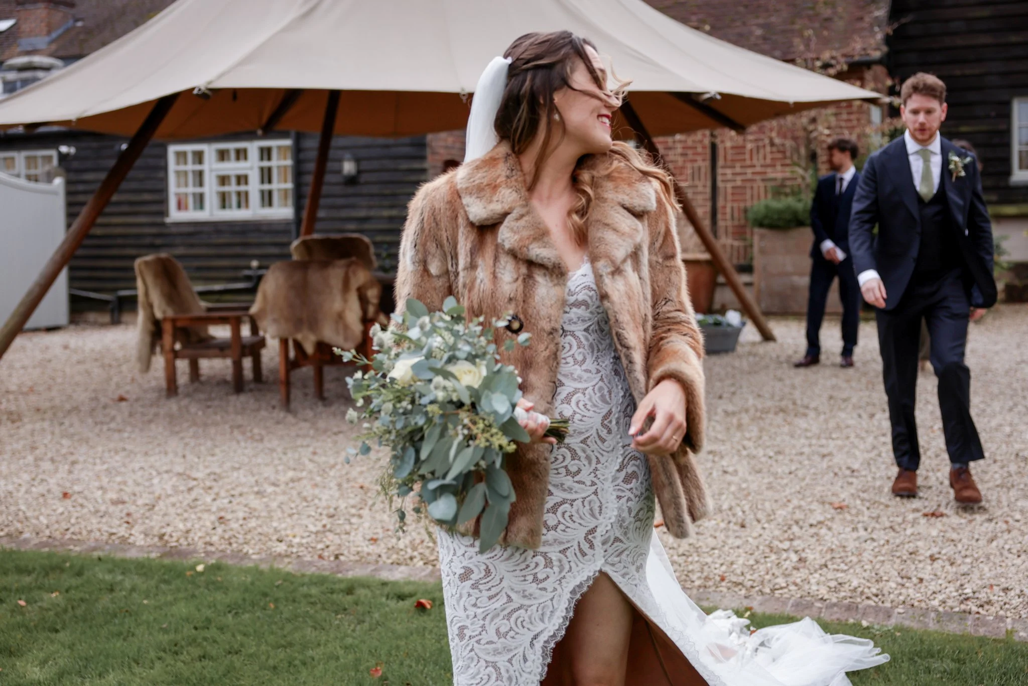 Bride in a white lace wedding dress and fur coat holding a bouquet, smiling, with multiple groomsmen and a wedding tent in the background.