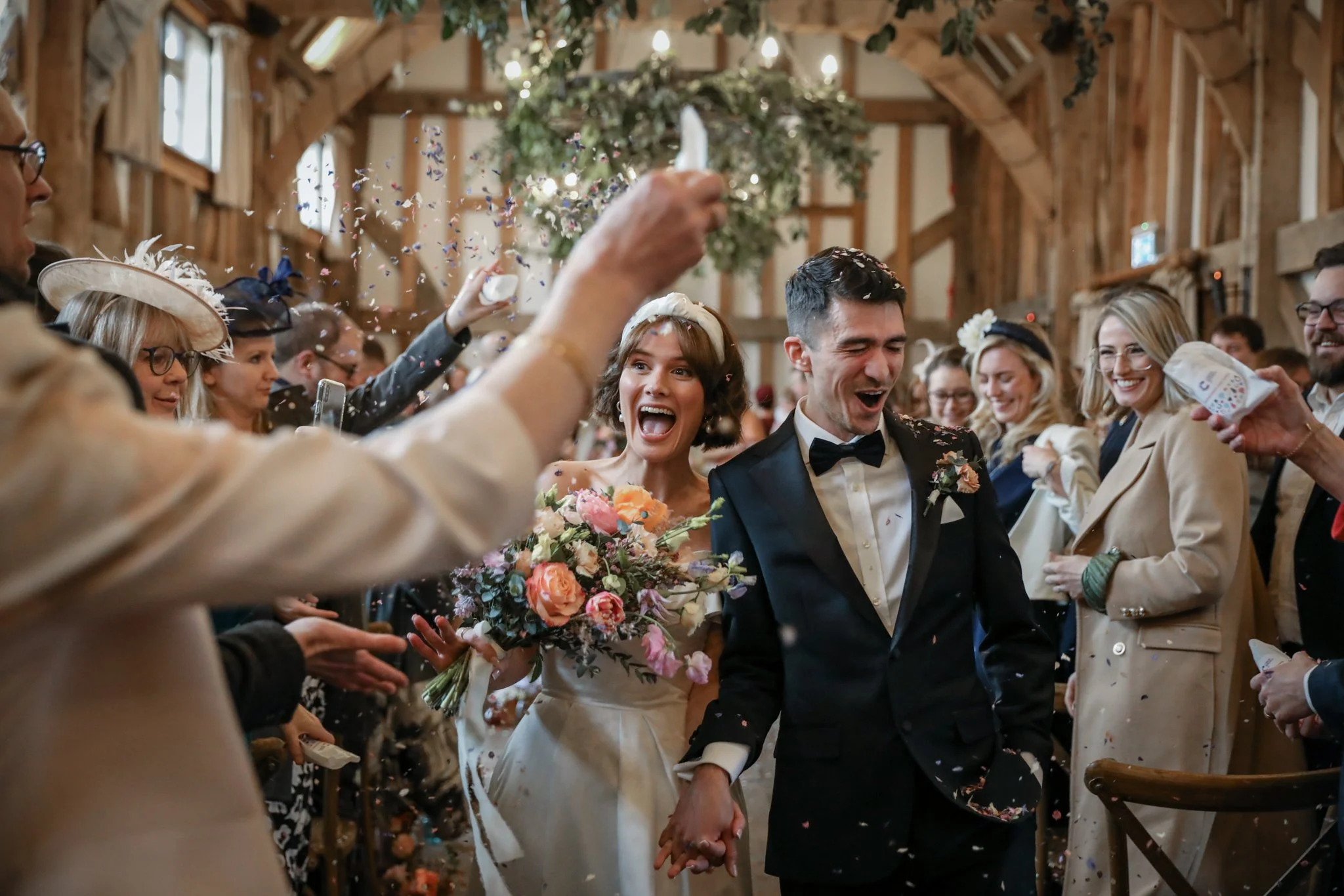 A newlywed couple is walking hand in hand down the aisle in a rustic barn, surrounded by friends and family celebrating with confetti and joyful expressions.