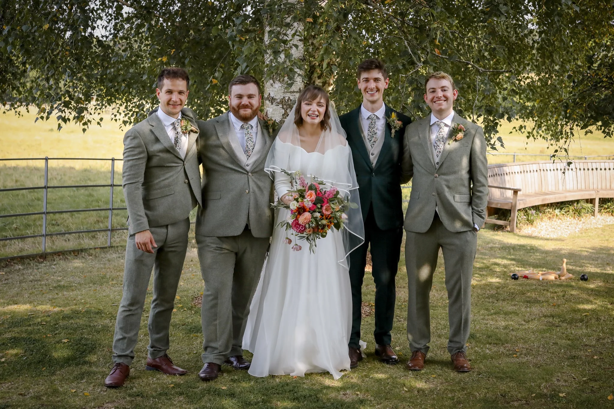 Group of five people dressed in wedding attire standing outdoors in front of a tree, smiling for a photo, with a park bench in the background.