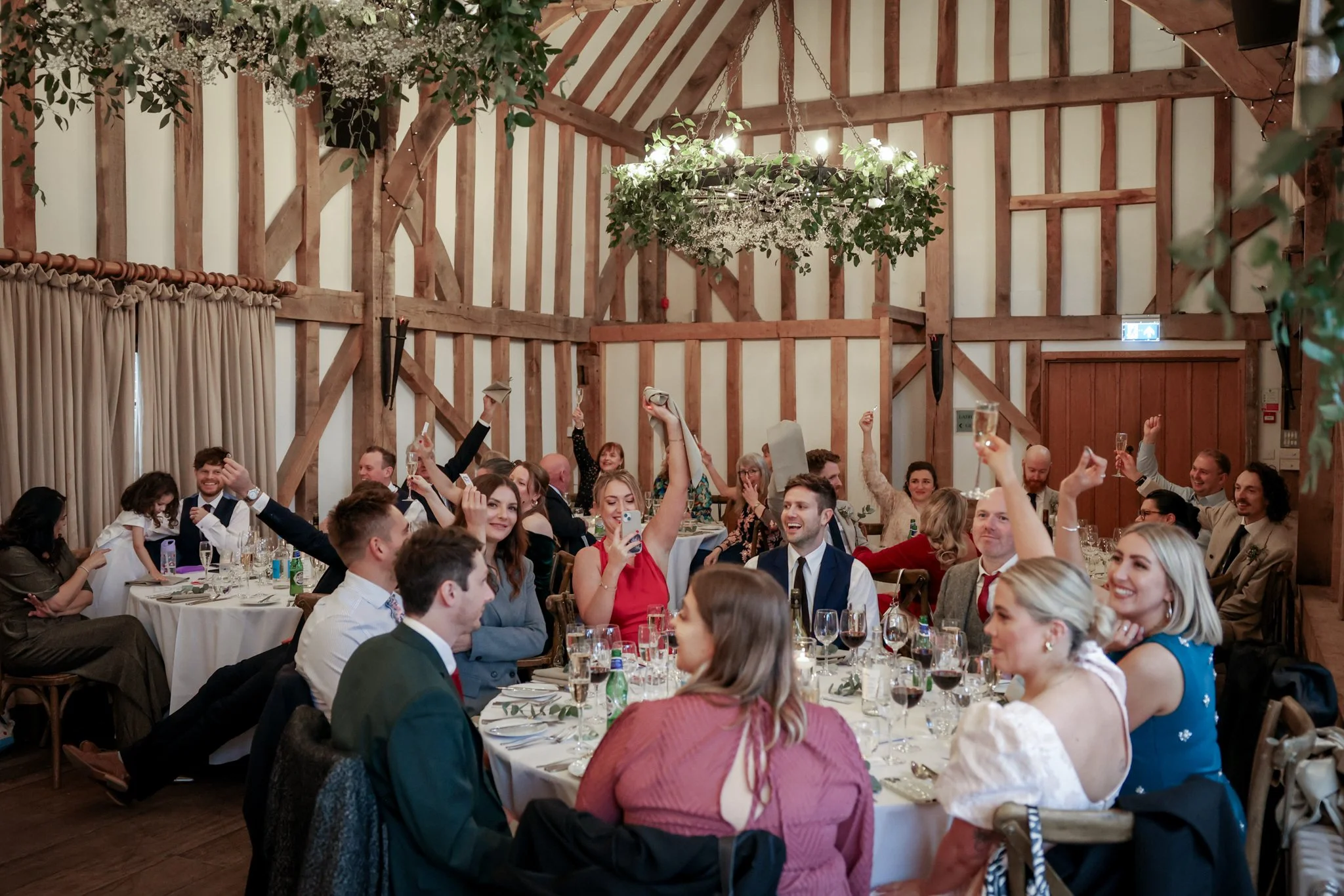 People gathered at a celebration or wedding reception in a rustic barn with wooden beams and a chandelier decorated with greenery and flowers.