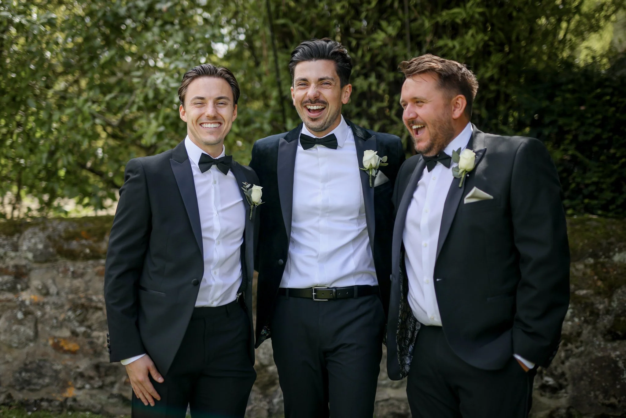 Three men in tuxedos smiling and laughing outdoors with greenery in the background.