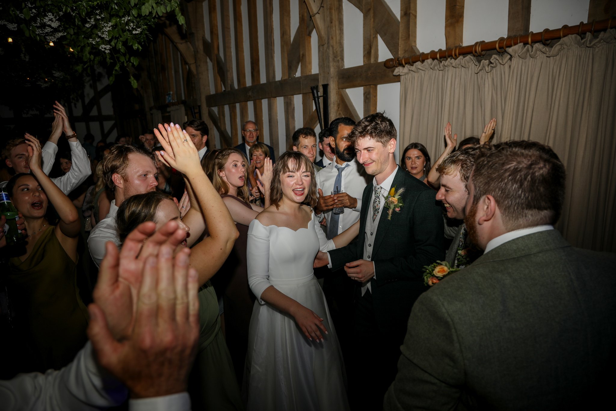A group of people at a wedding celebration, with the bride and groom at the center, smiling and enjoying the moment, surrounded by friends and family in a rustic venue.