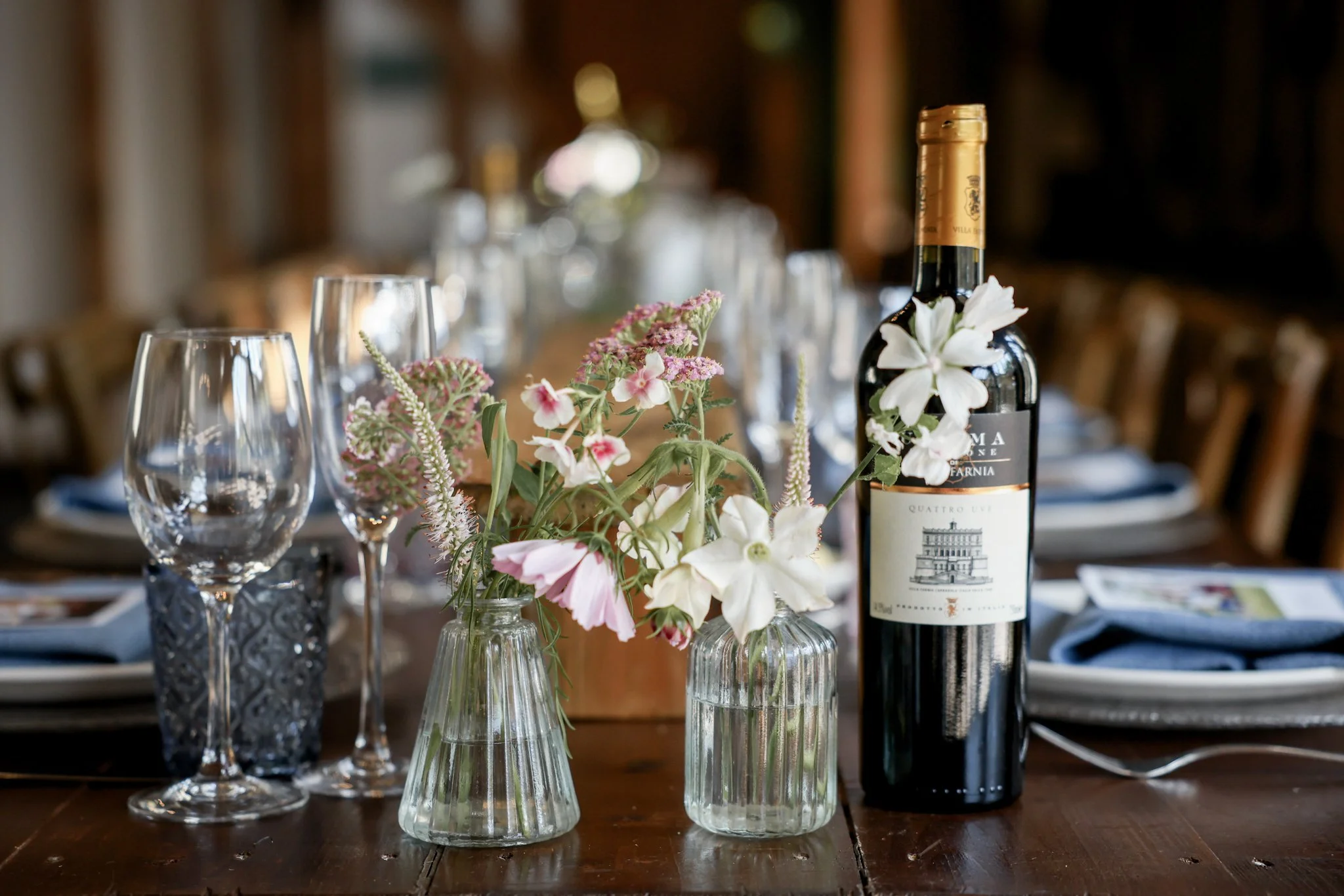 A dining table decorated with pink and white flowers in glass vases, a bottle of Italian wine, and empty wine glasses, set in a cozy, warmly lit room.