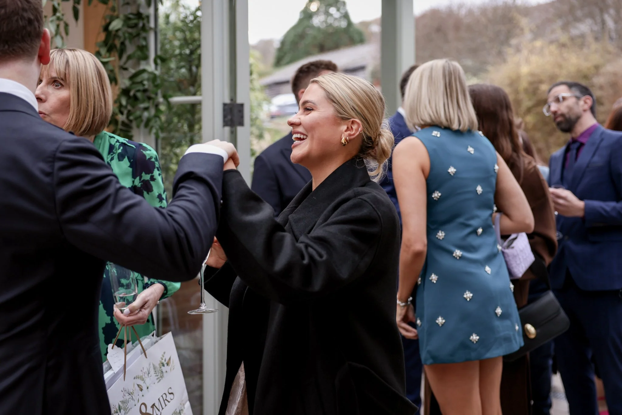 People dressed in formal attire, smiling and socializing at an outdoor event with a glass of champagne, in a garden or conservatory setting.
