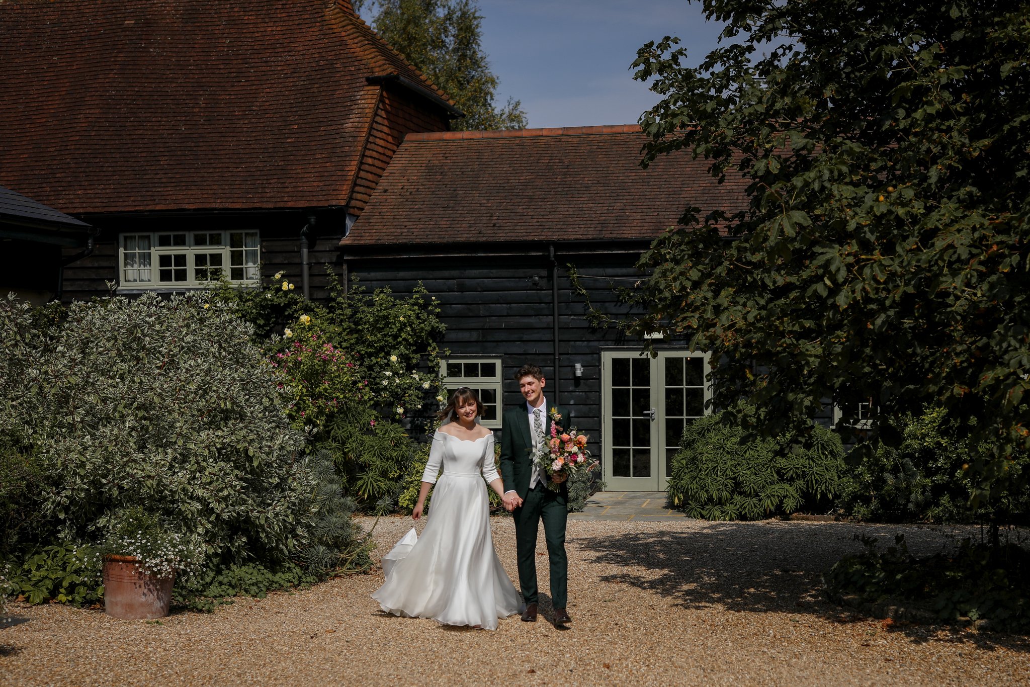 A bride and groom smiling and holding hands outdoors in front of a dark wooden house with a red tile roof, surrounded by greenery, during daytime.