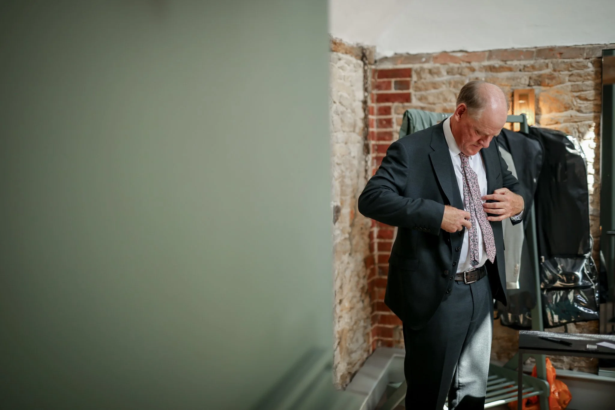 A man in a suit and tie adjusting his shirt in a room with exposed brick walls.