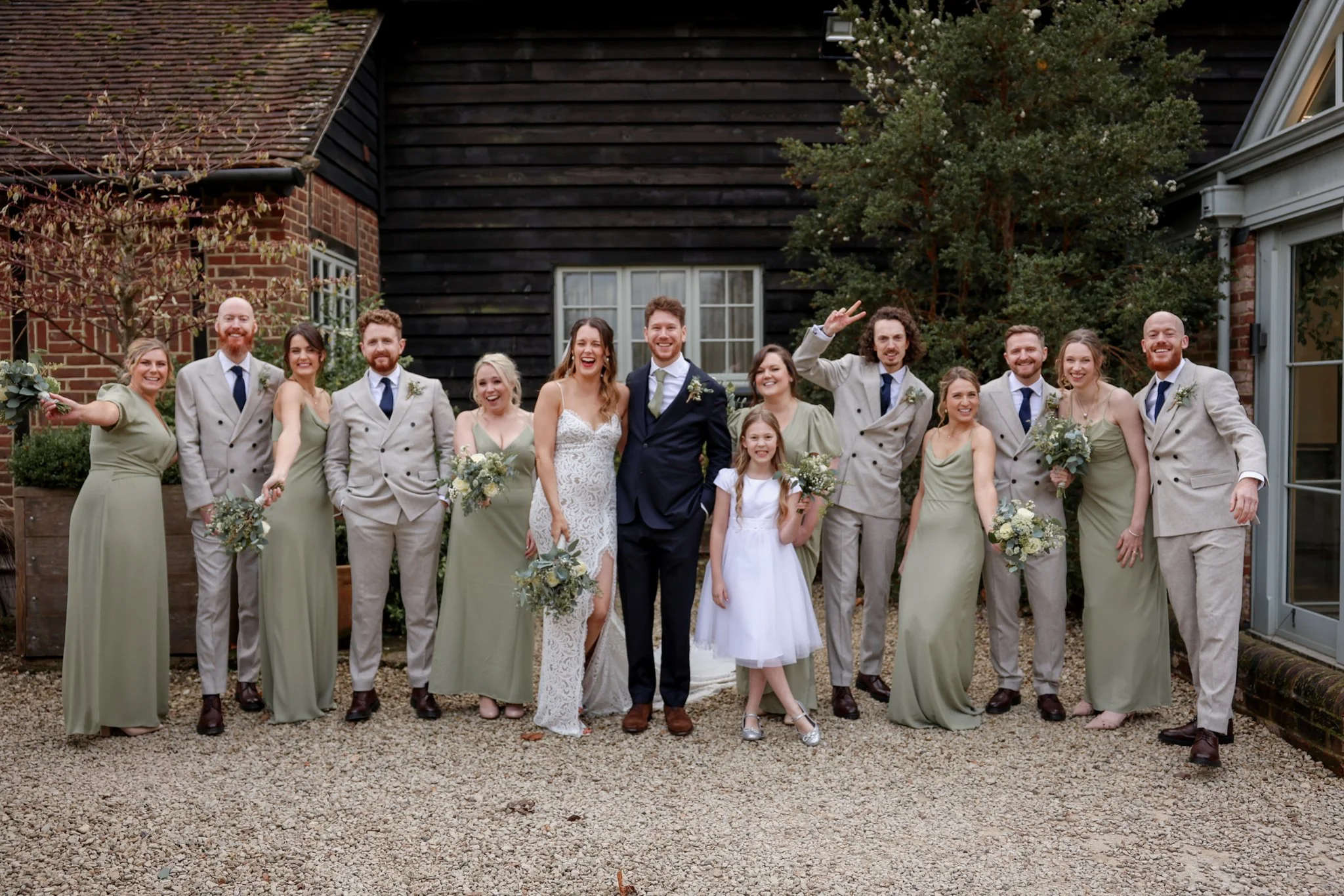 A group of sixteen people, including men, women, and children, dressed in wedding attire, posing outside a house with brick and black wooden siding. The bride is wearing a white lace dress, and the groom is in a dark suit. The others are wearing ligh