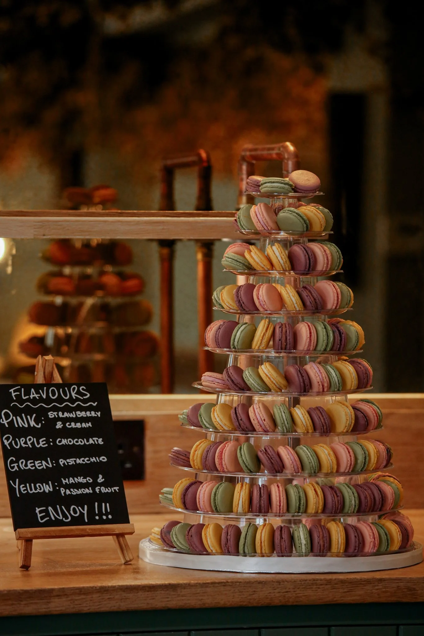 Colorful macarons displayed on a multi-tiered tray in a bakery or cafe setting, with a chalkboard sign listing flavors and a blurred background of a warm, rustic interior.
