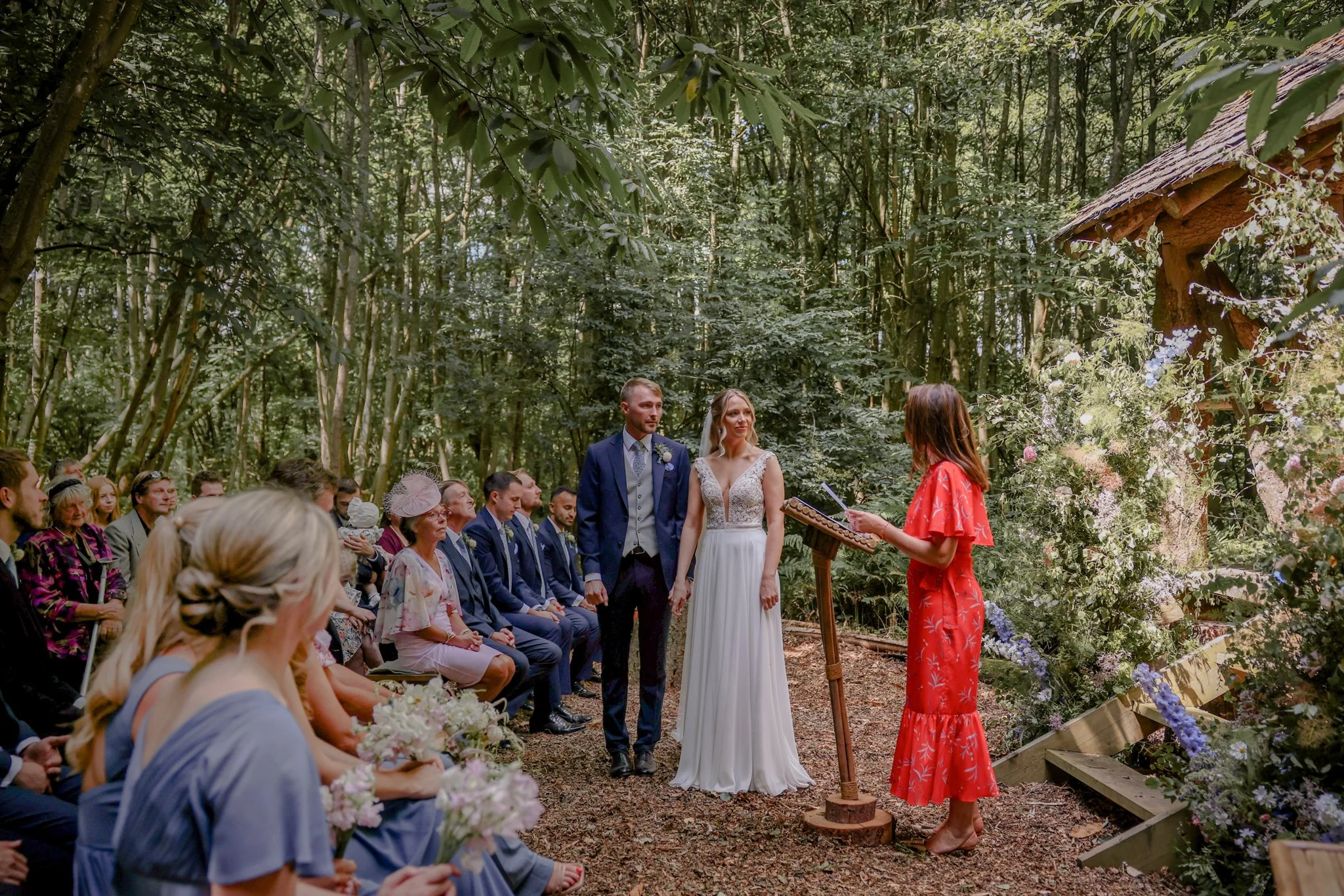 A wedding ceremony taking place outdoors in a forest, with a bride and groom standing before a woman in a red dress reading from a book, surrounded by seated guests and floral decorations.