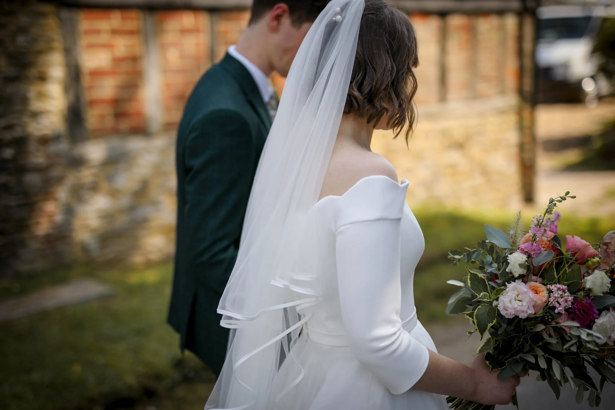 A bride and groom standing outdoors during a wedding, with the bride holding a bouquet of flowers and the groom dressed in a suit. The bride's dress is white with a veil, and the groom's head is close to her, both with their heads bowed.